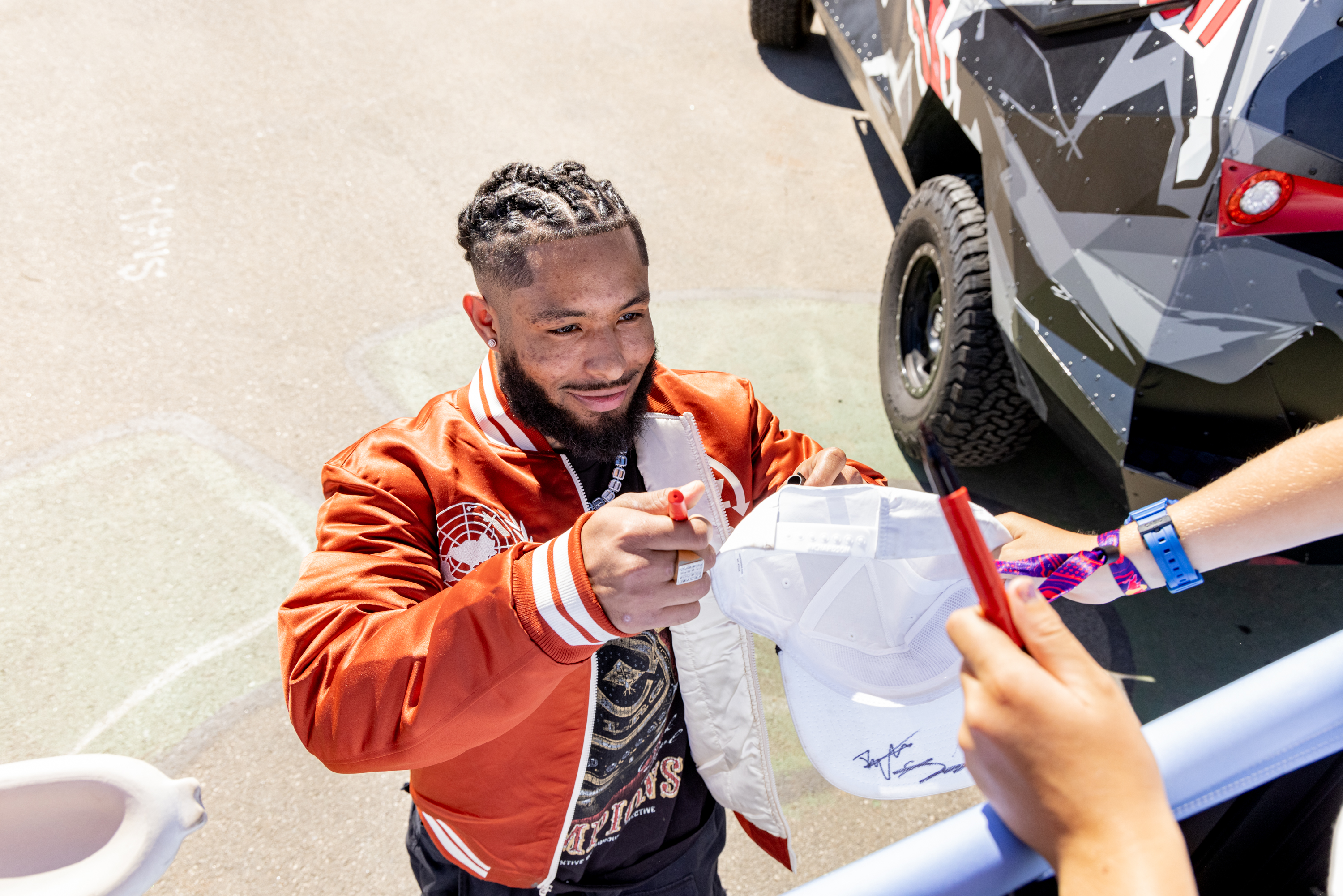 A man in an orange jacket signs a white cap for someone. He smiles, holding a red marker. A vehicle with graphic designs is parked nearby.