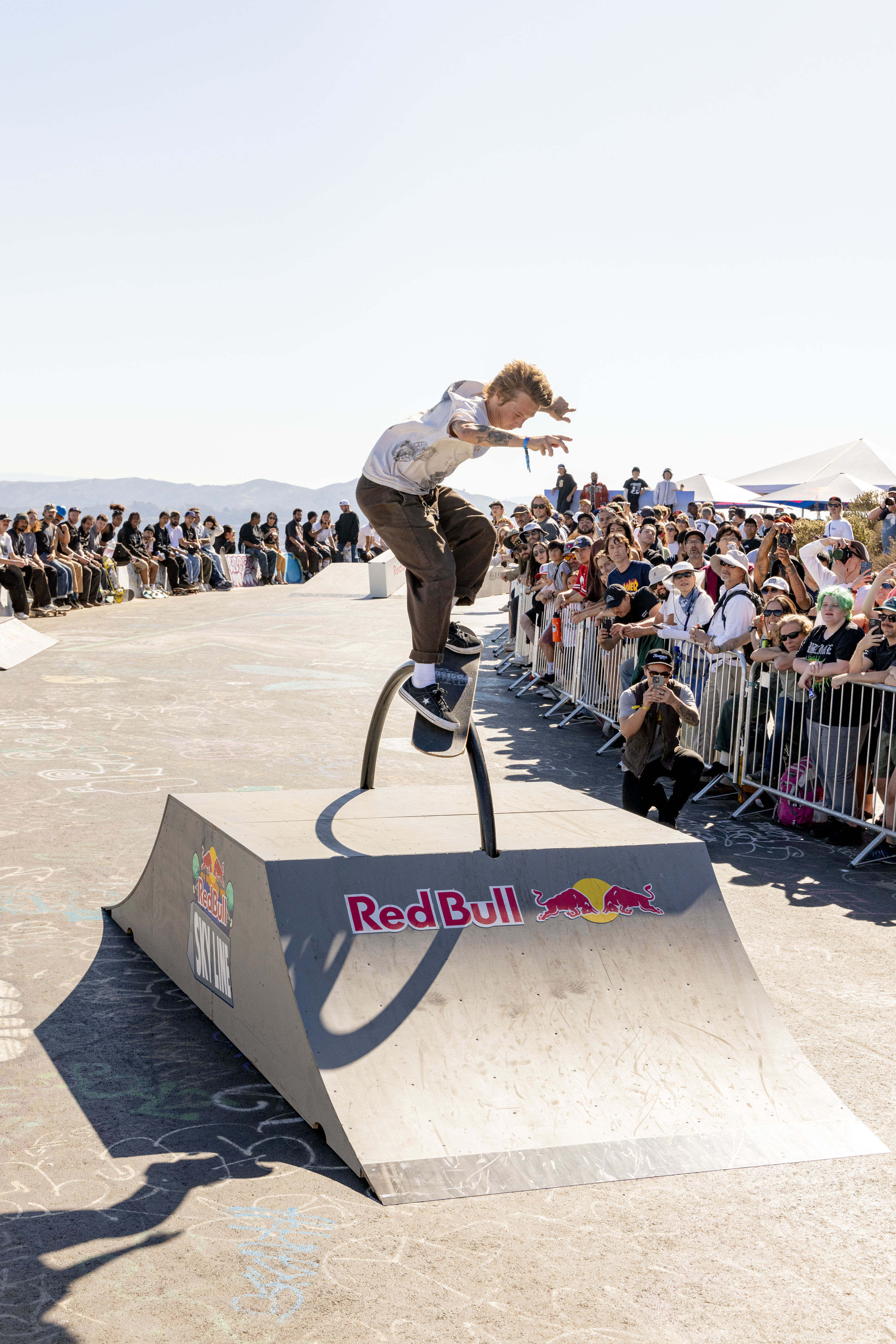 A skateboarder performs a trick on a curved rail ramp, surrounded by a large crowd of spectators. The ramp displays Red Bull branding.