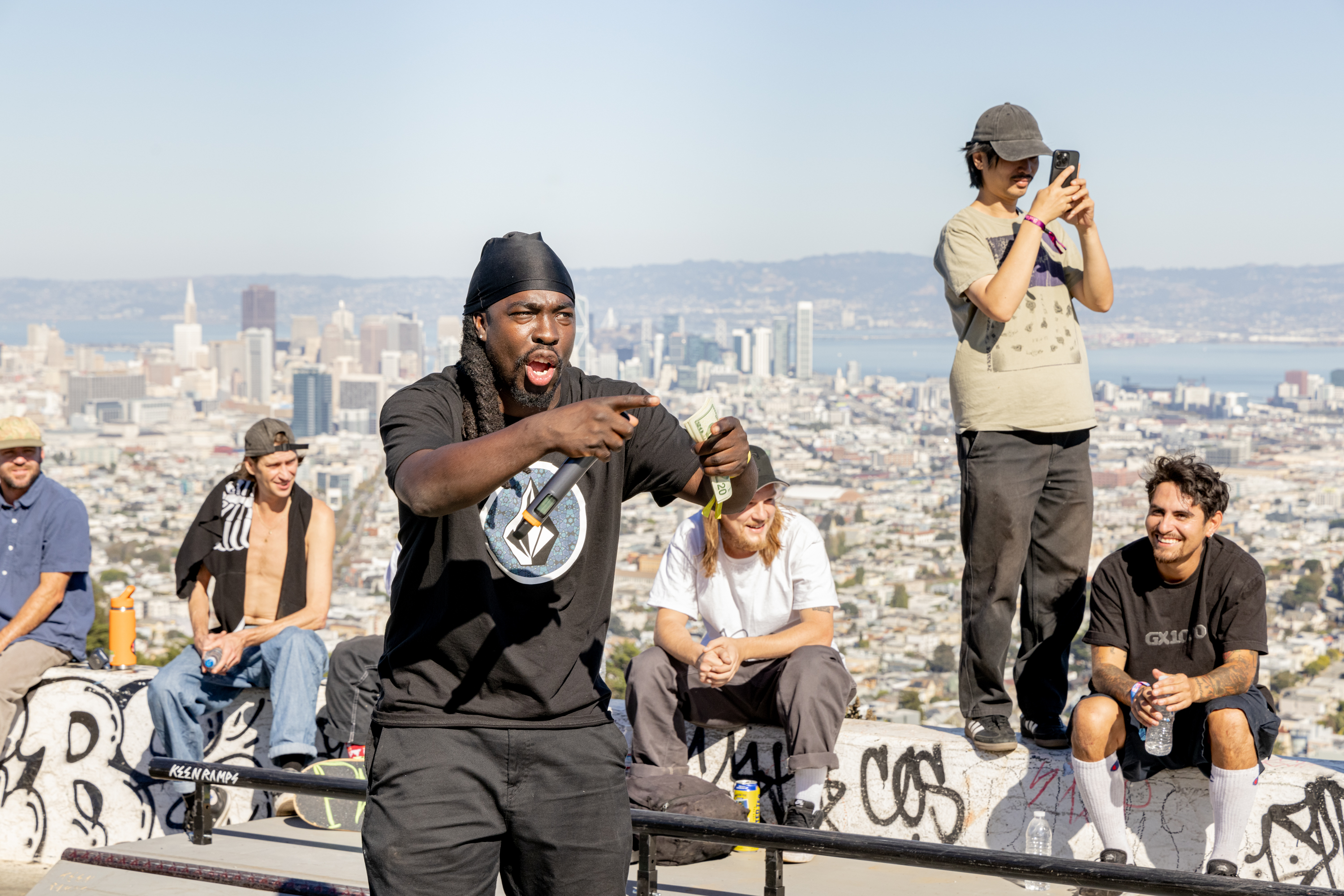 A group of people are at a graffiti-covered skate park with a city skyline in the background. One person is excitedly holding a microphone and cash.