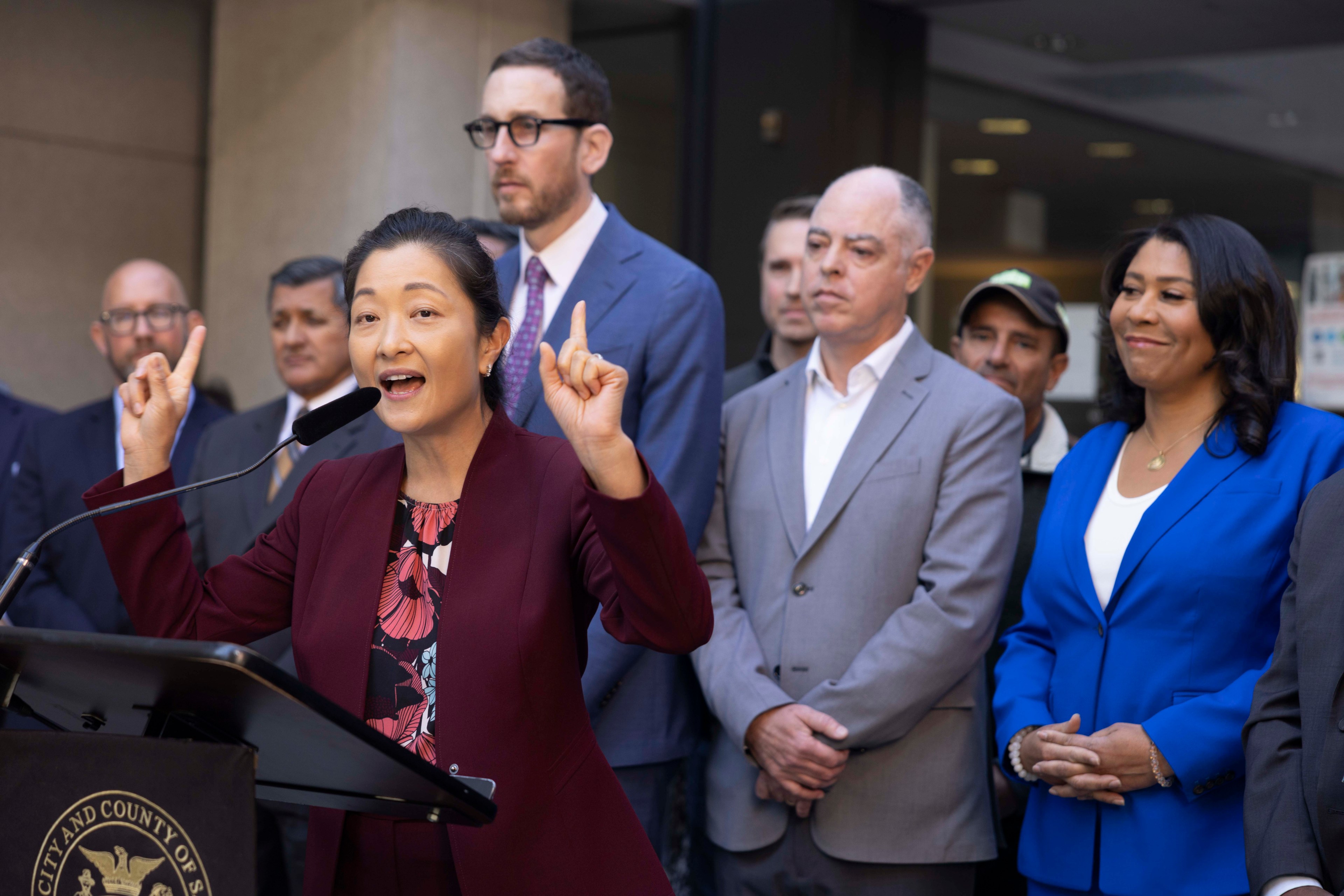 A woman in a maroon suit speaks passionately at a podium, surrounded by several attentive people in suits and a woman in a blue blazer.