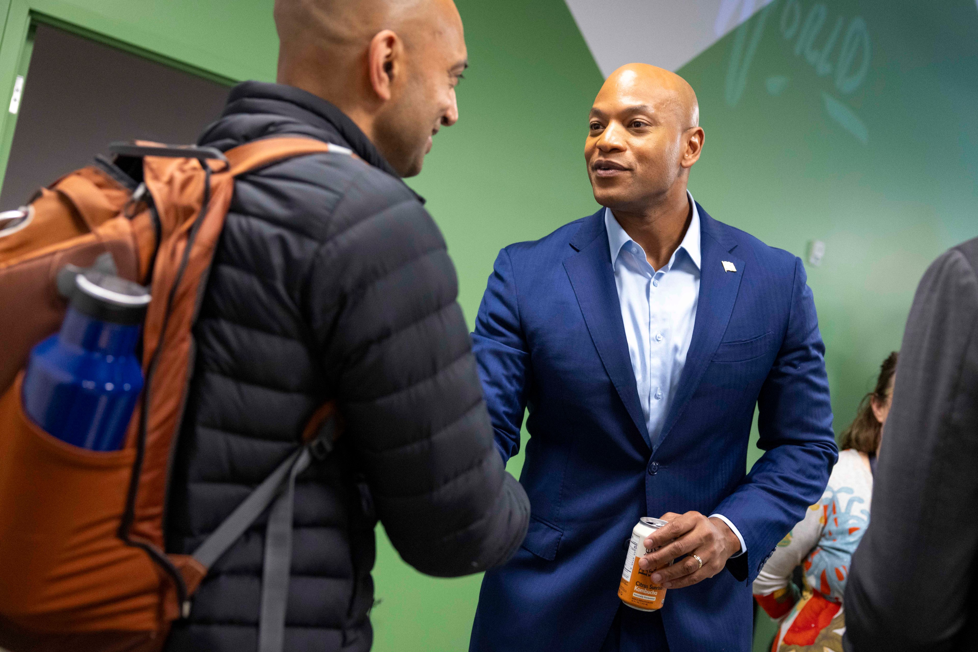 A man in a blue suit shakes hands with another man wearing a backpack and holding a blue water bottle, against a green wall.