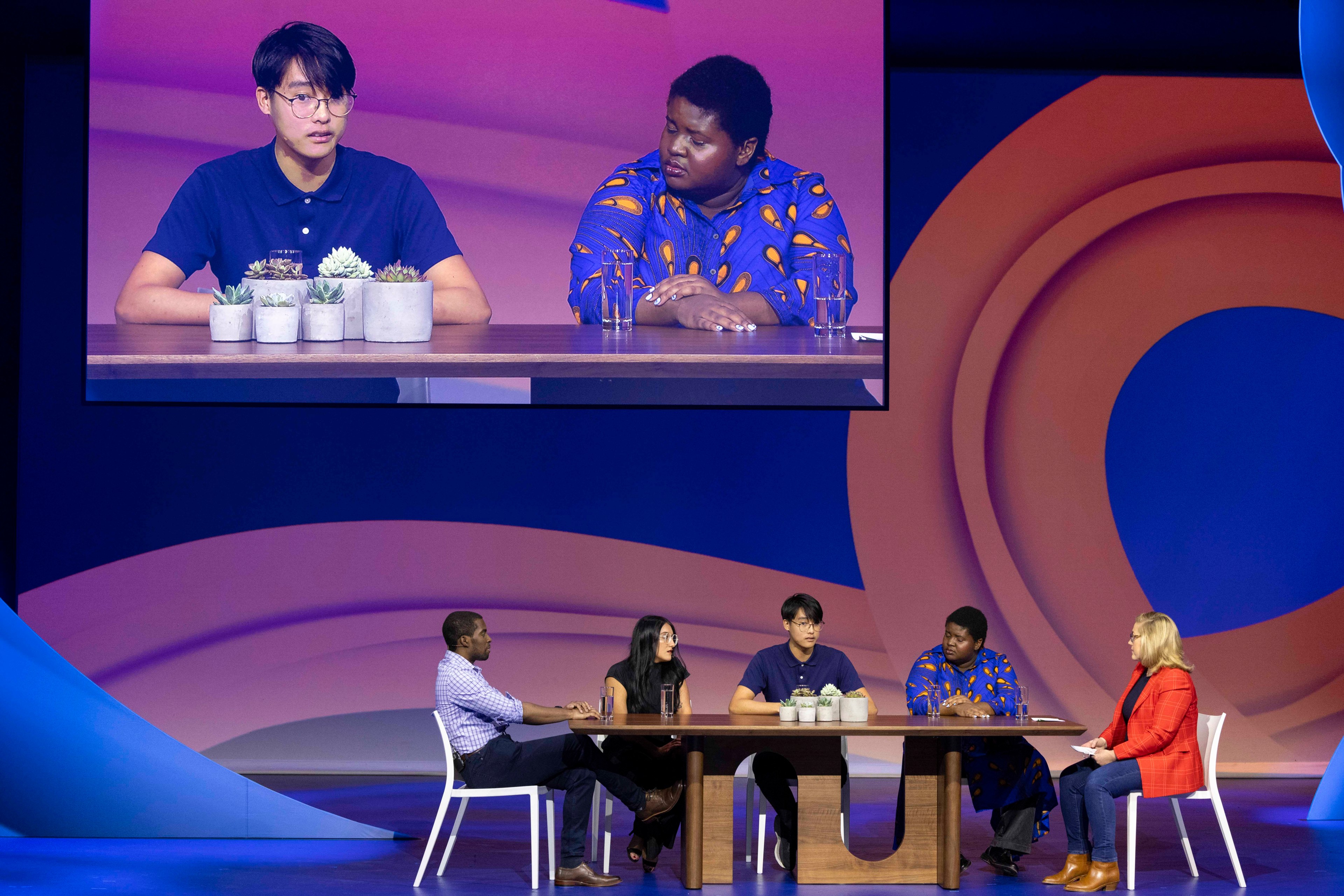 Five people sit at a table on stage, engaged in a discussion. A large screen above displays a close-up of two of them, with potted plants in view.