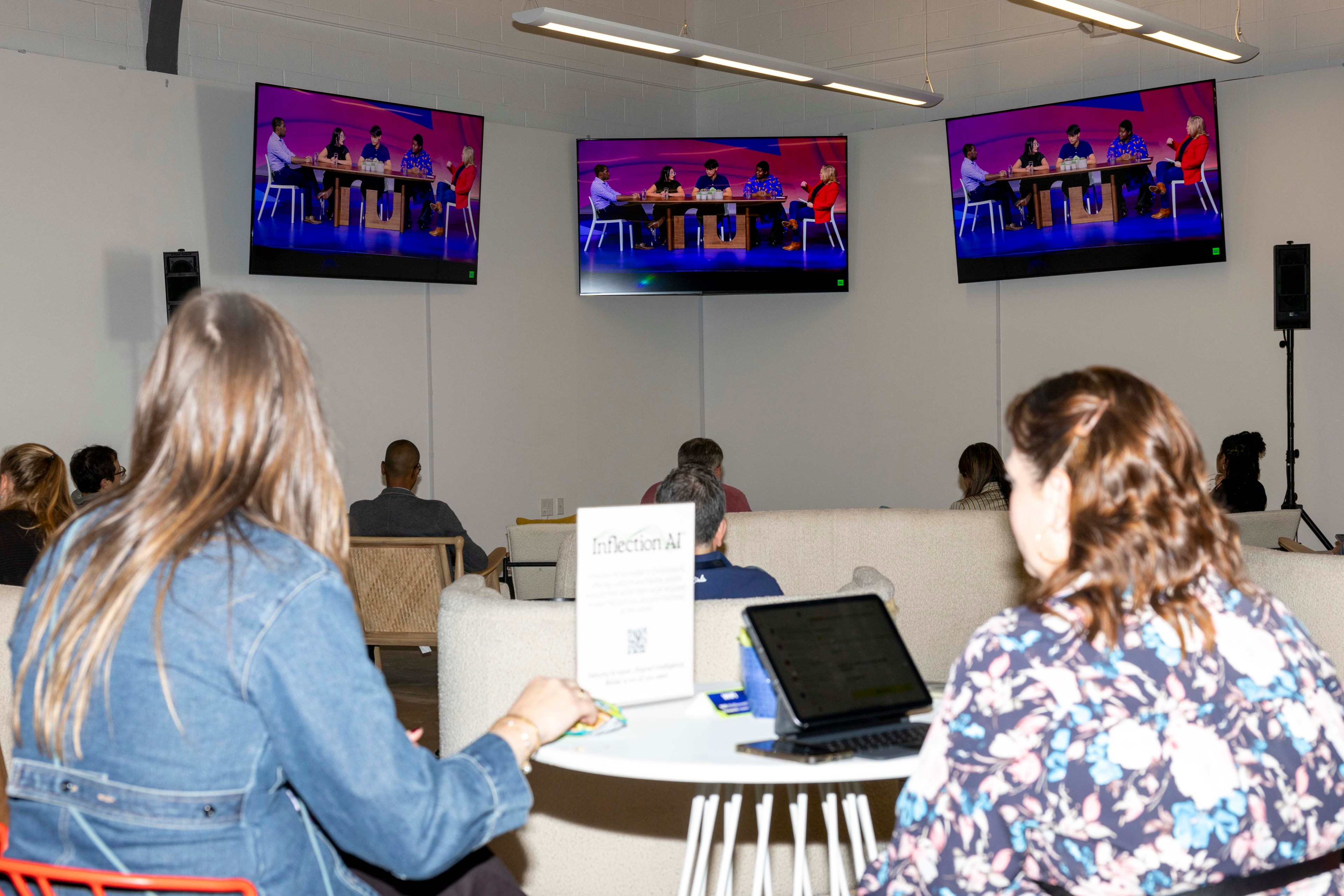 People are seated in a room watching a panel discussion on three large screens. A &quot;Inflection AI&quot; sign is visible on a table with a laptop.