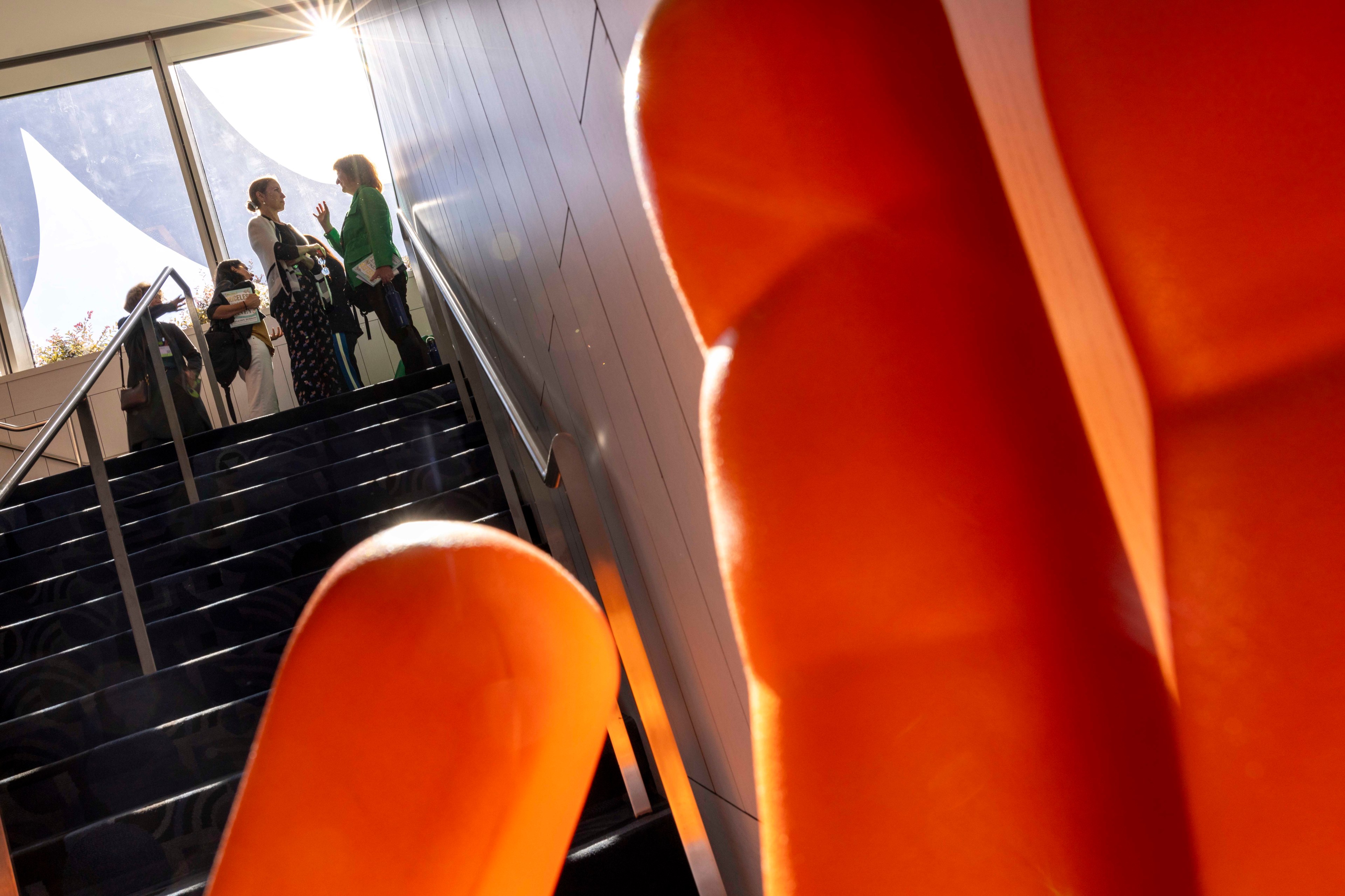 Two people are talking at the top of a staircase bathed in sunlight, with orange abstract shapes in the foreground and bright windows in the background.