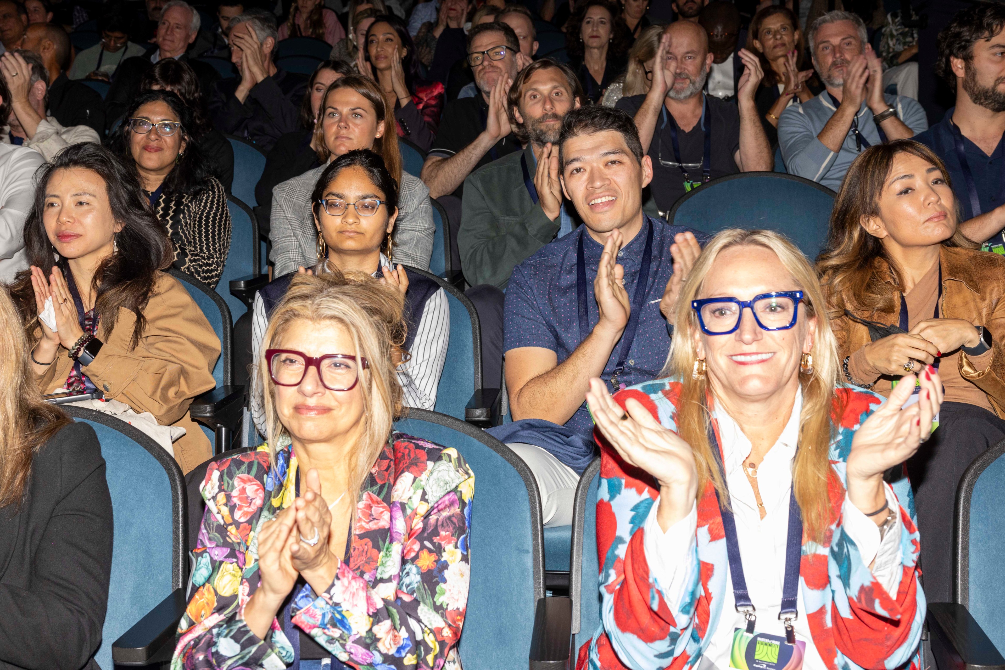 A diverse group of people is sitting in an auditorium, clapping and smiling. They appear engaged and enthusiastic, with some wearing colorful clothing and glasses.