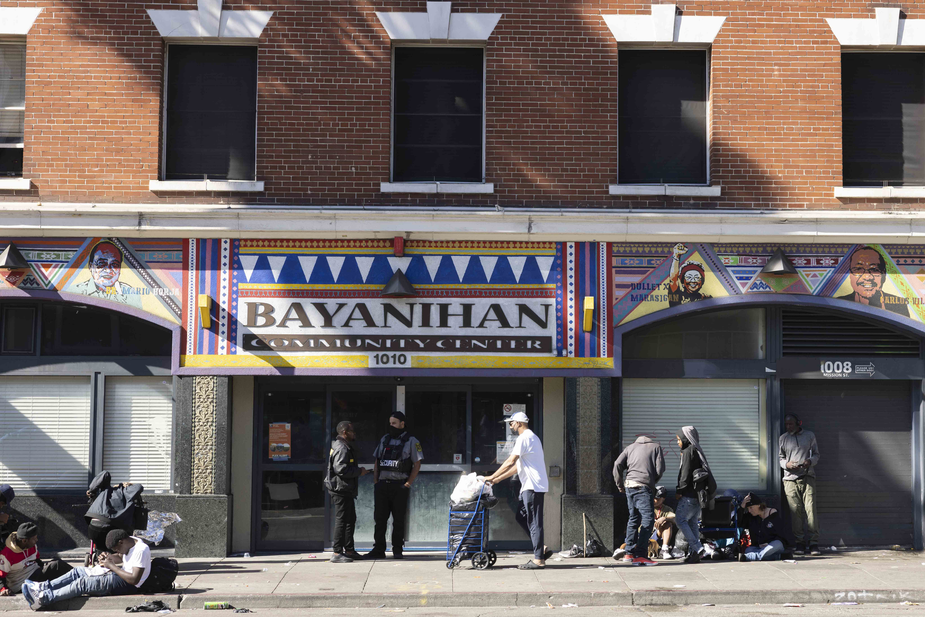 The image depicts the Bayanihan Community Center with a colorful facade featuring artwork. Several people are gathered outside, including individuals with bags and carts.