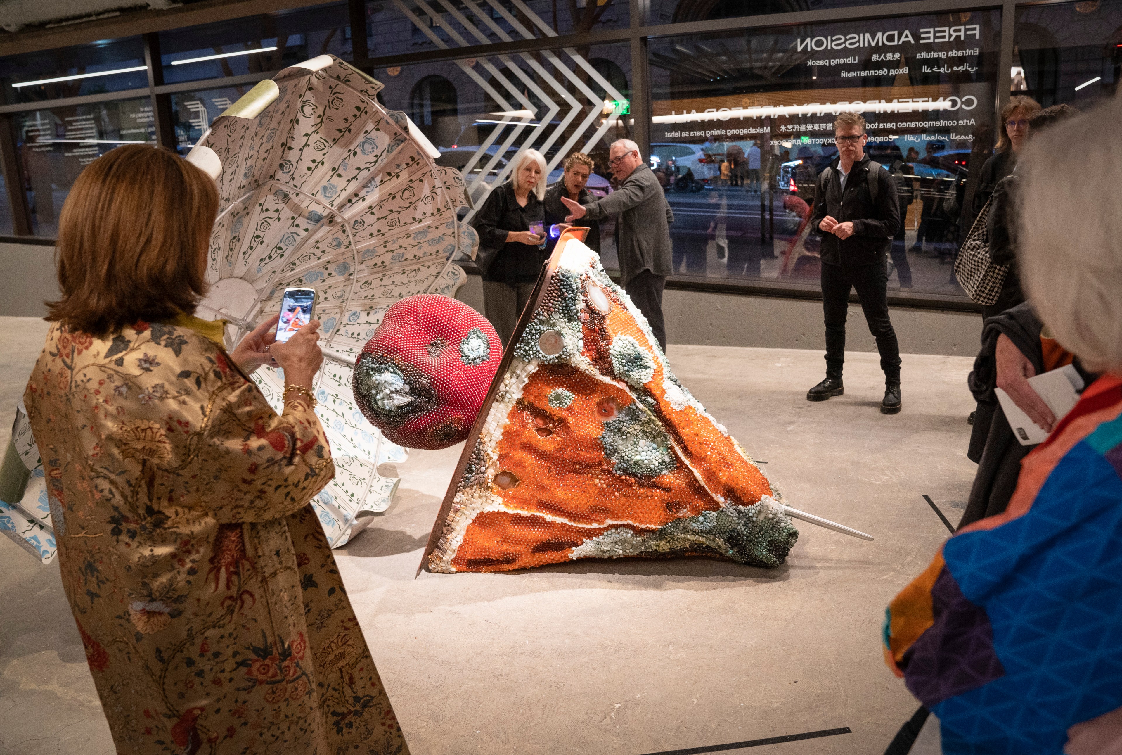 A group of people are viewing a colorful, abstract sculpture that resembles oversized food items. One person is taking a photo with their phone.