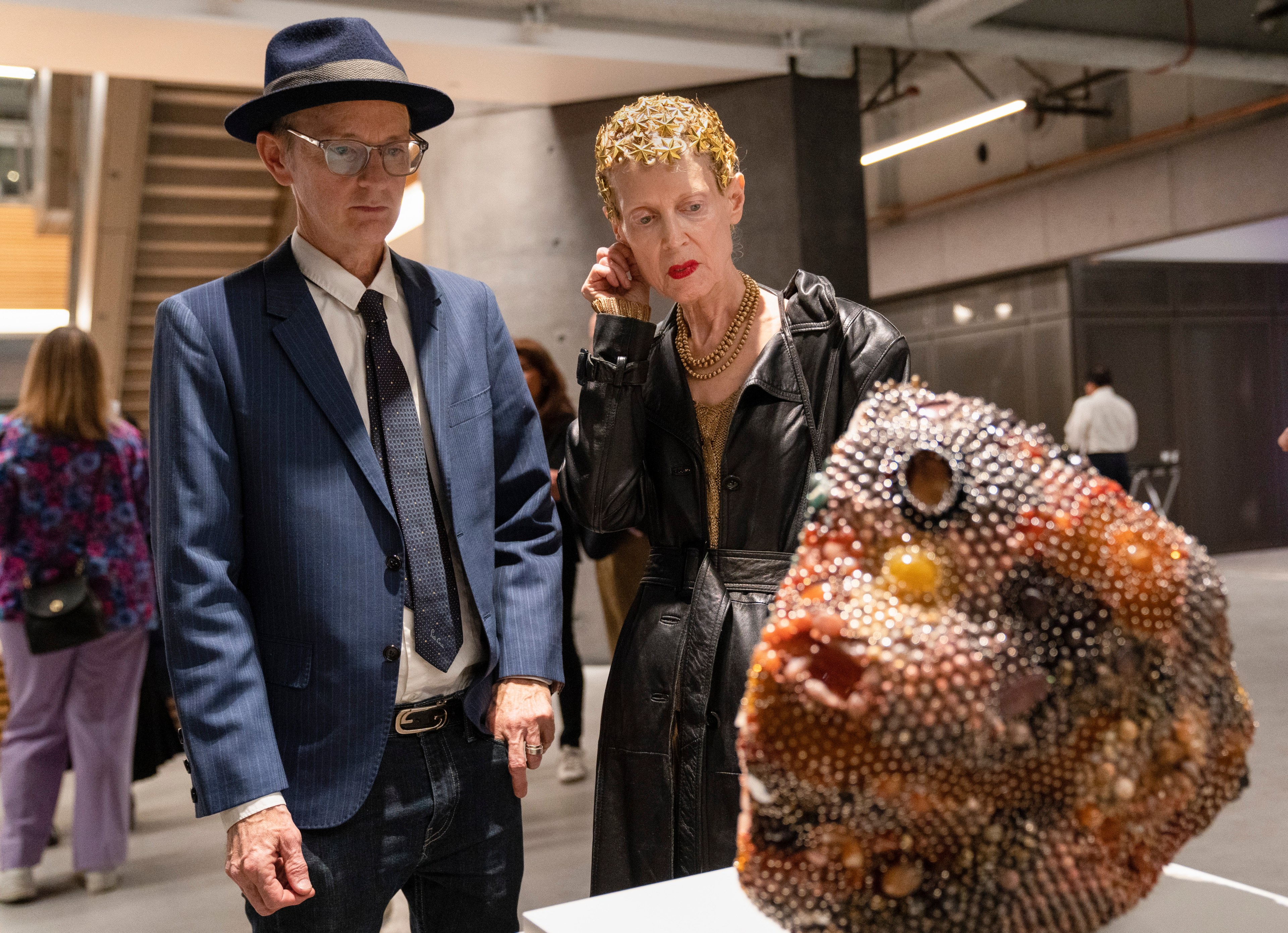 A man in a blue suit and hat stands next to a woman in a black coat, gazing at a textured, colorful sculpture. The background shows an indoor art gallery space.