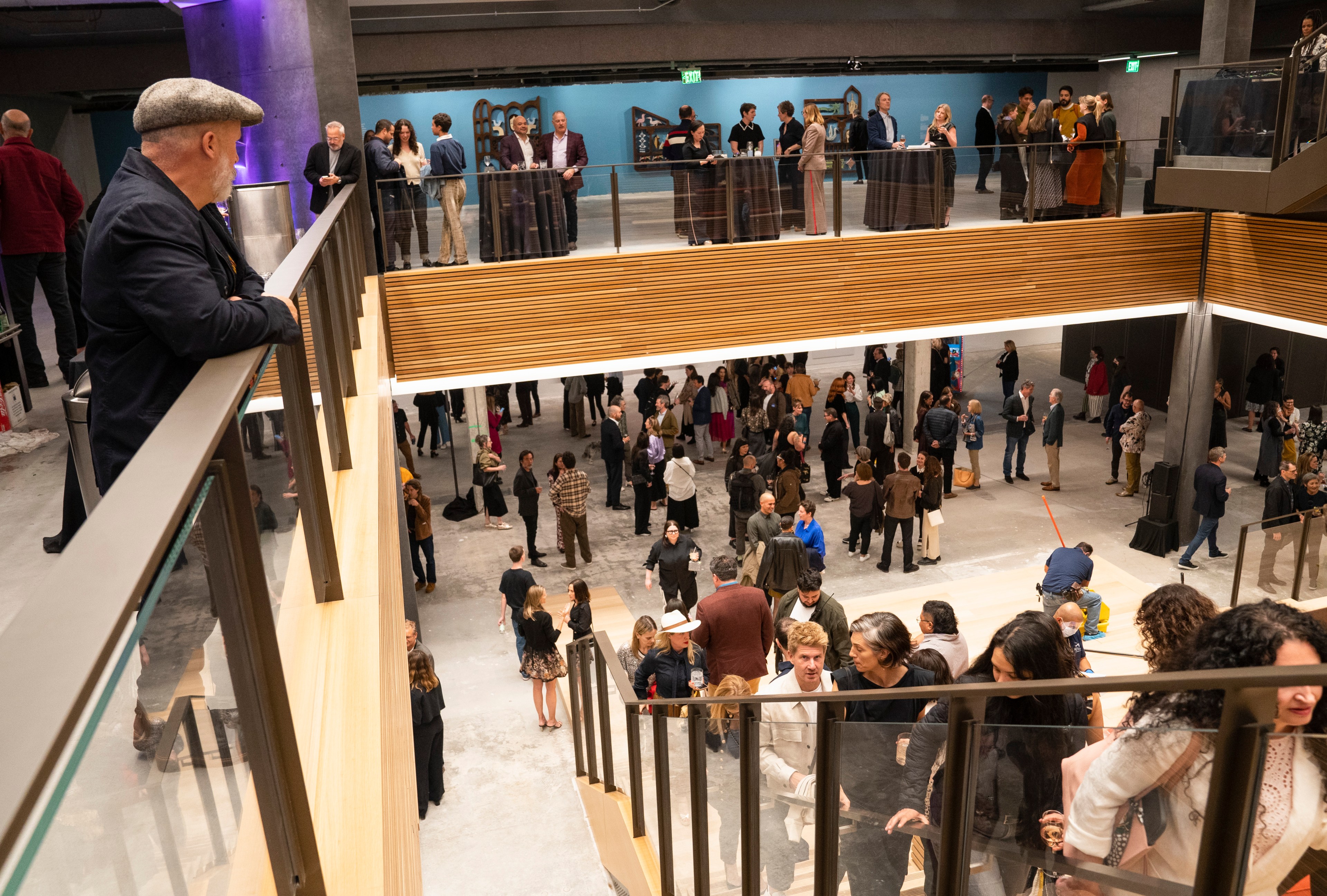 A crowded indoor event with people on two levels, featuring a modern space with a wooden staircase and glass railings, and vibrant conversations all around.