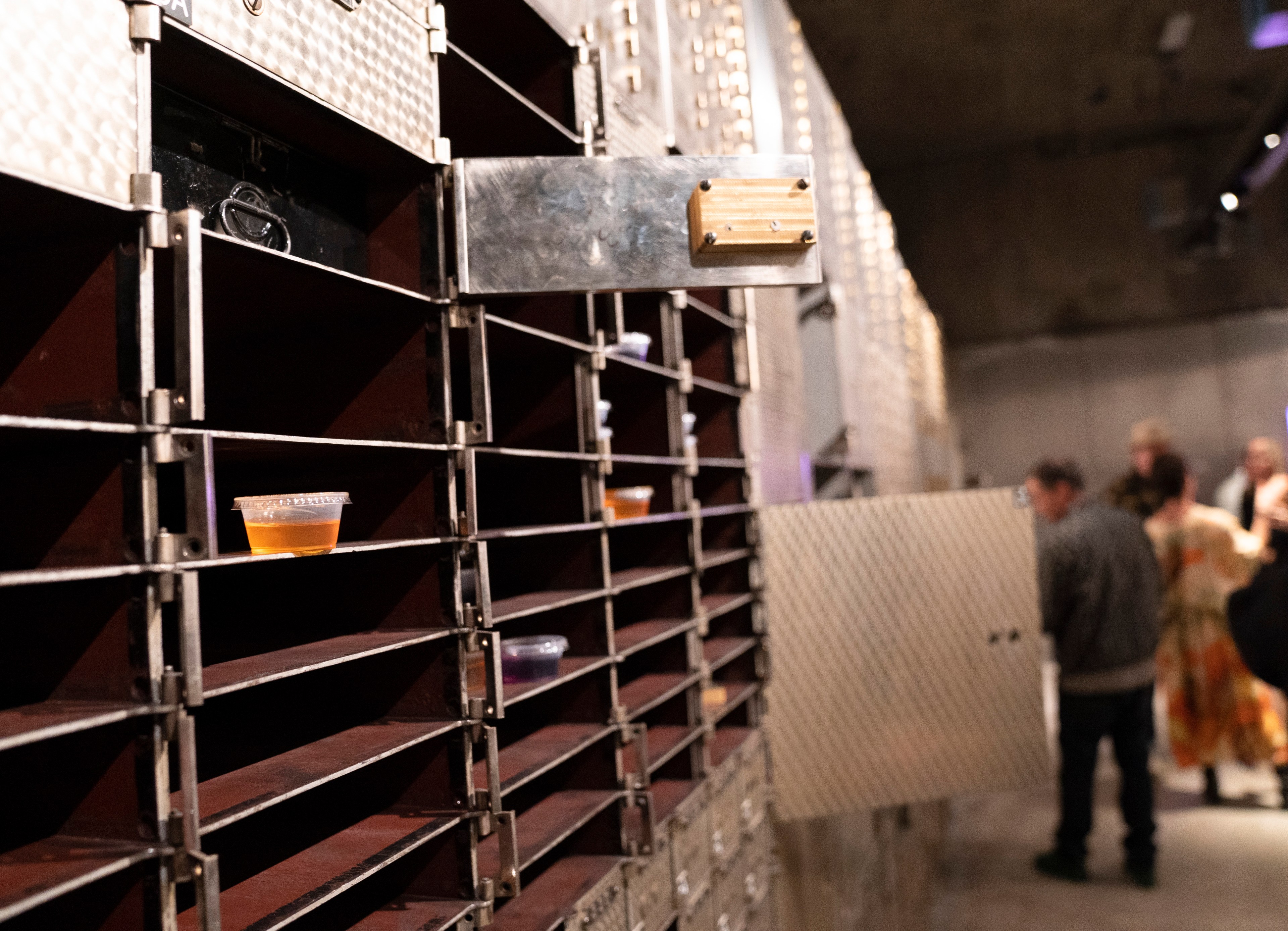 The image shows industrial-style shelves with small cups of liquid on them. In the background, a group of people stand, slightly out of focus.