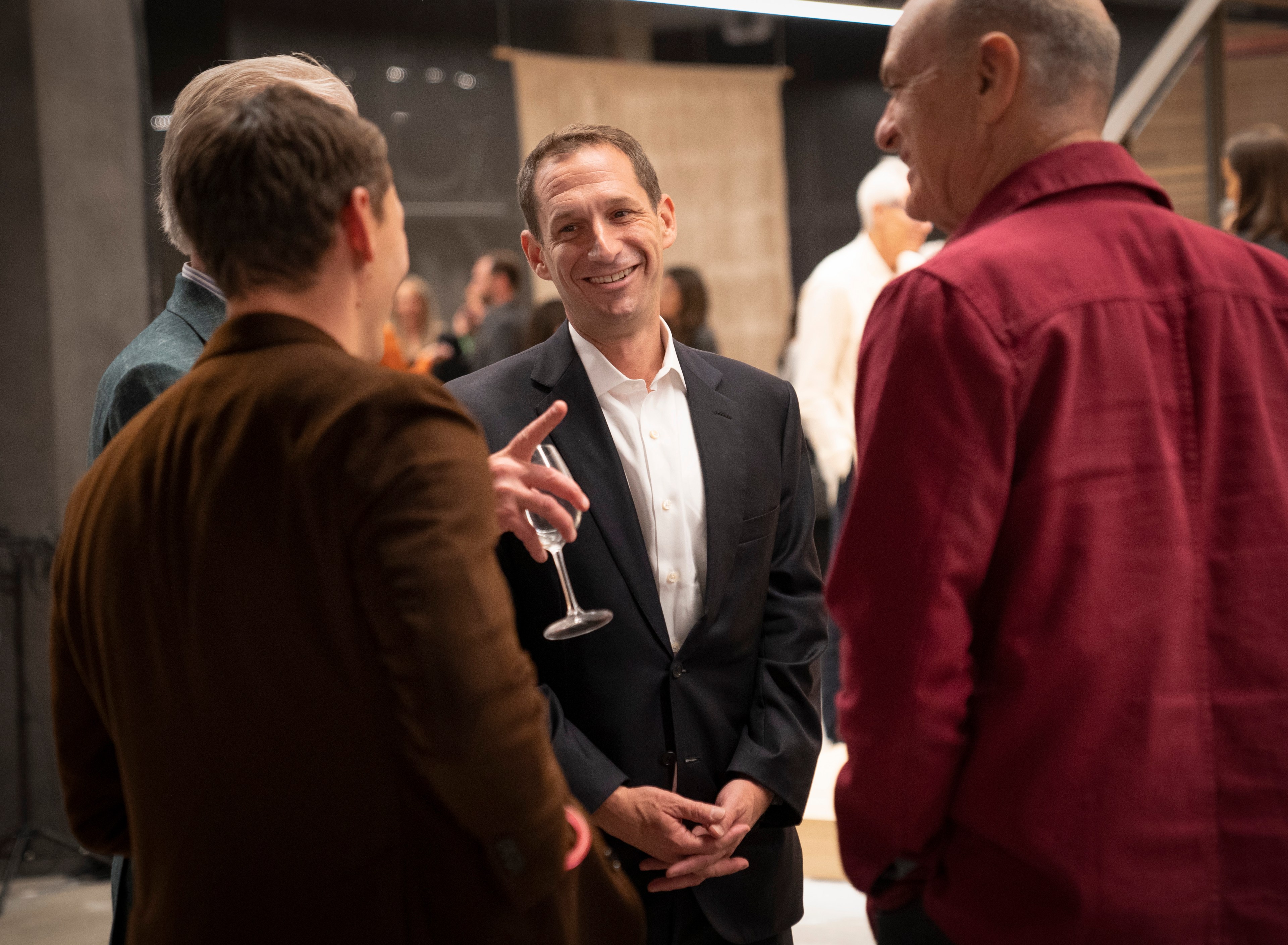 A man in a suit smiles warmly in a group conversation at a social event. He stands among casually dressed people, one holding a champagne glass.