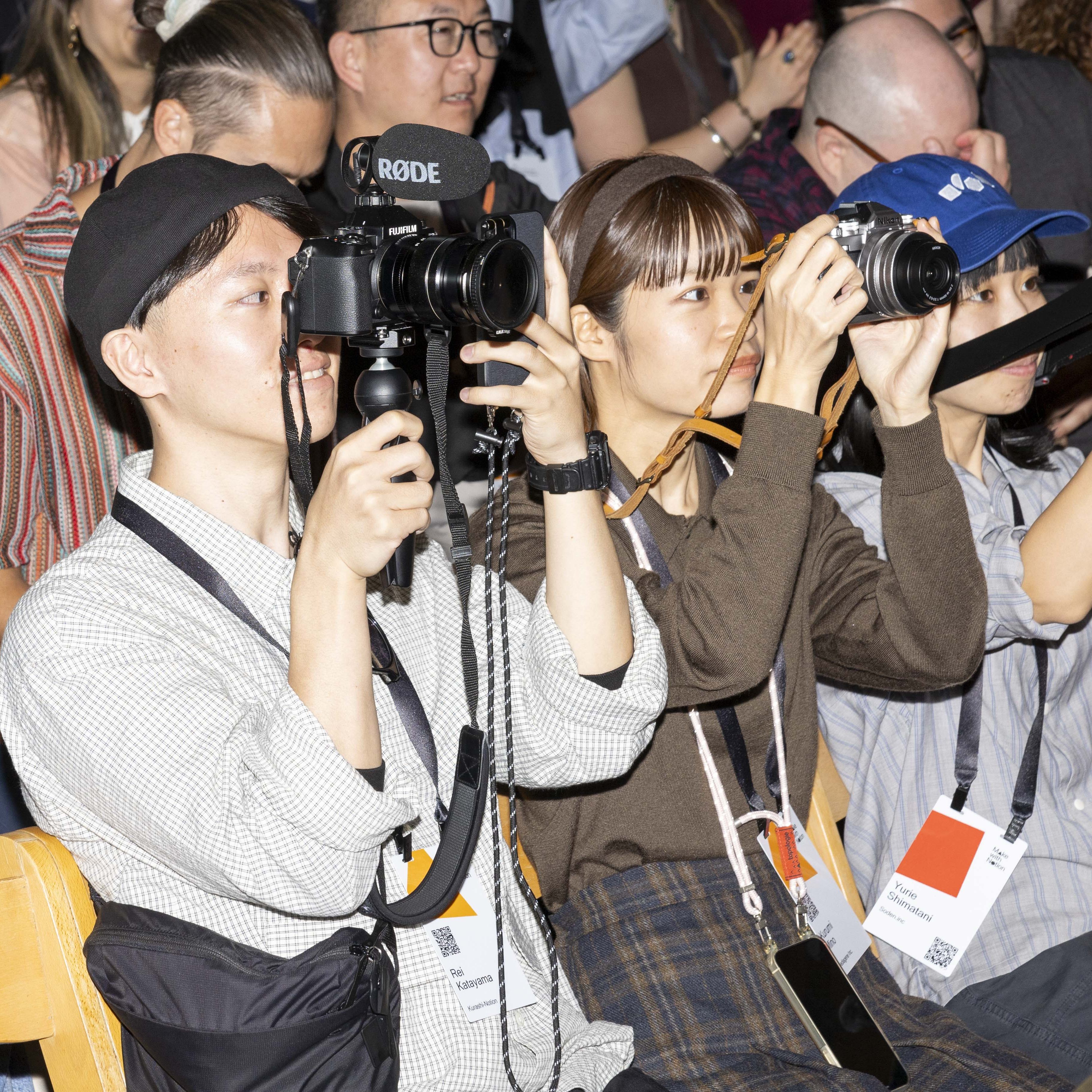 People are sitting in rows, holding cameras and taking photos, wearing name tags around their necks. They are focused intently on the event in front of them.