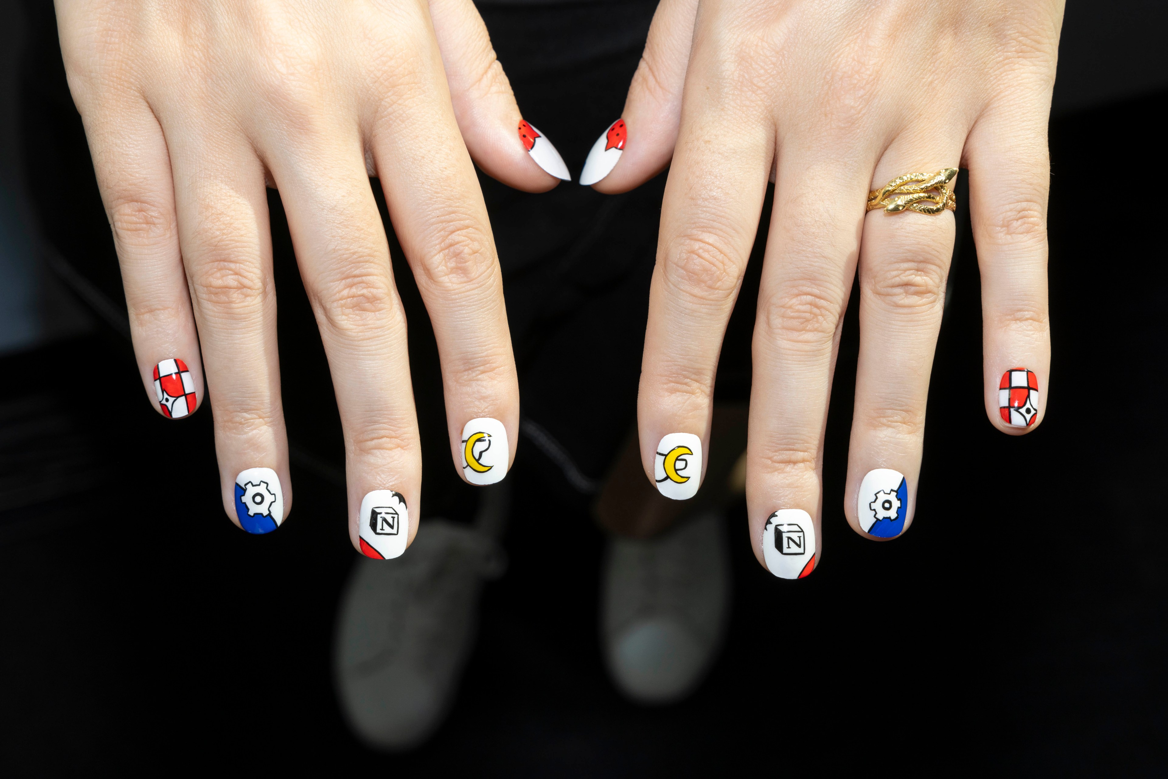 Two hands with nails painted in white feature various colorful designs, including moons, shields, and gears. A gold ring adorns one finger.