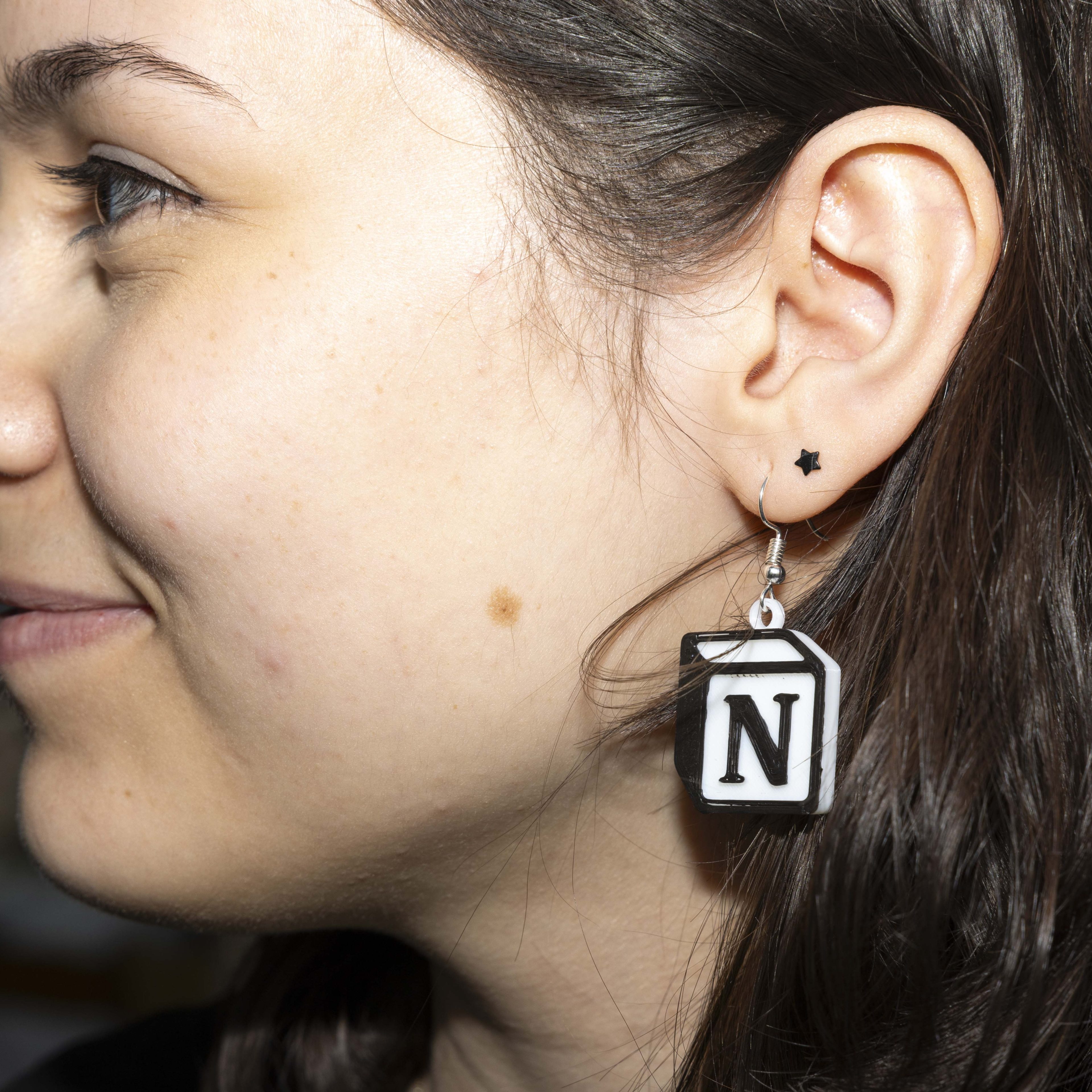 A person is wearing a large dangling earring in the shape of a black and white letter &quot;N&quot; document icon. They also have a small star-shaped earring.