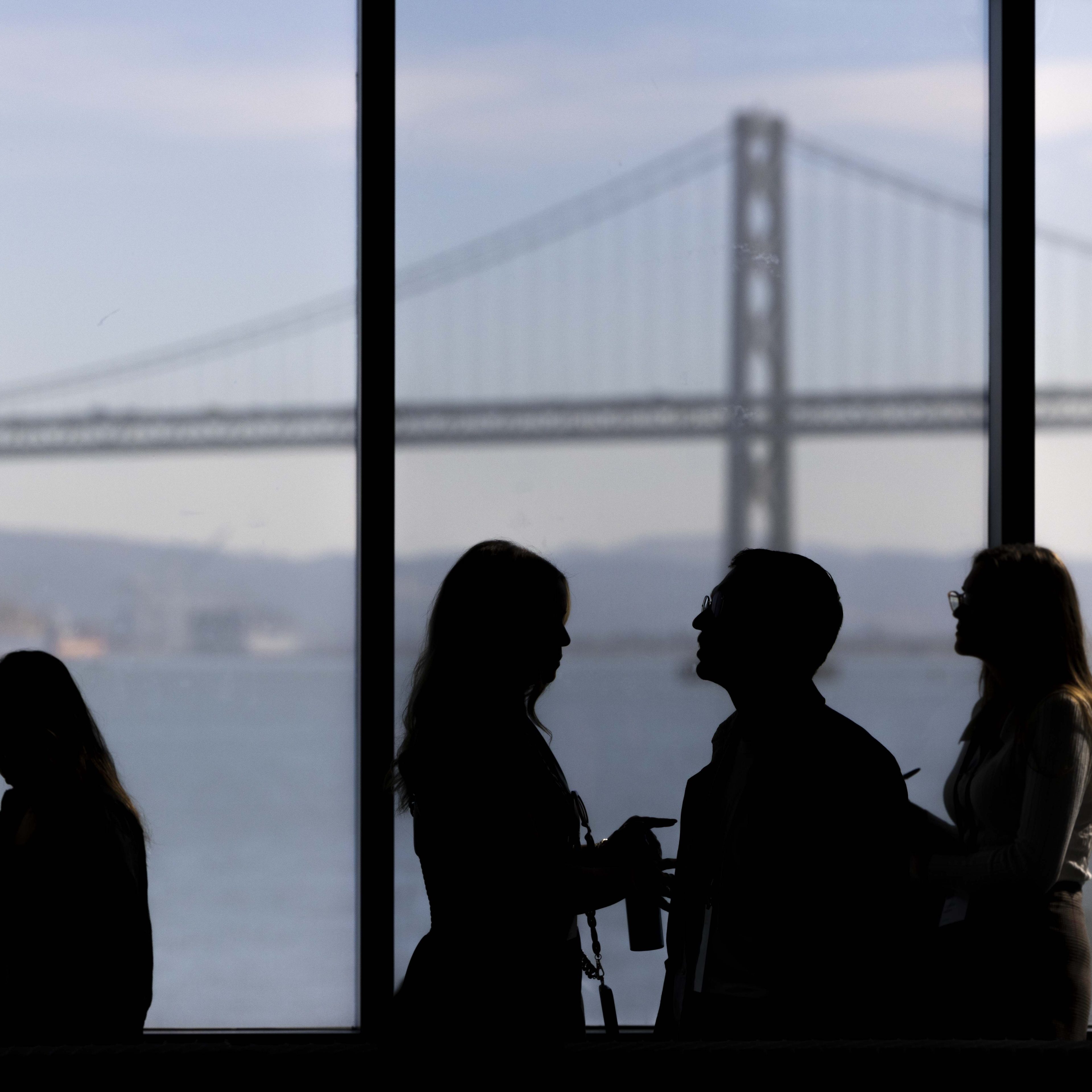 Silhouettes of four people stand by a window, with a large suspension bridge and a body of water visible in the background.