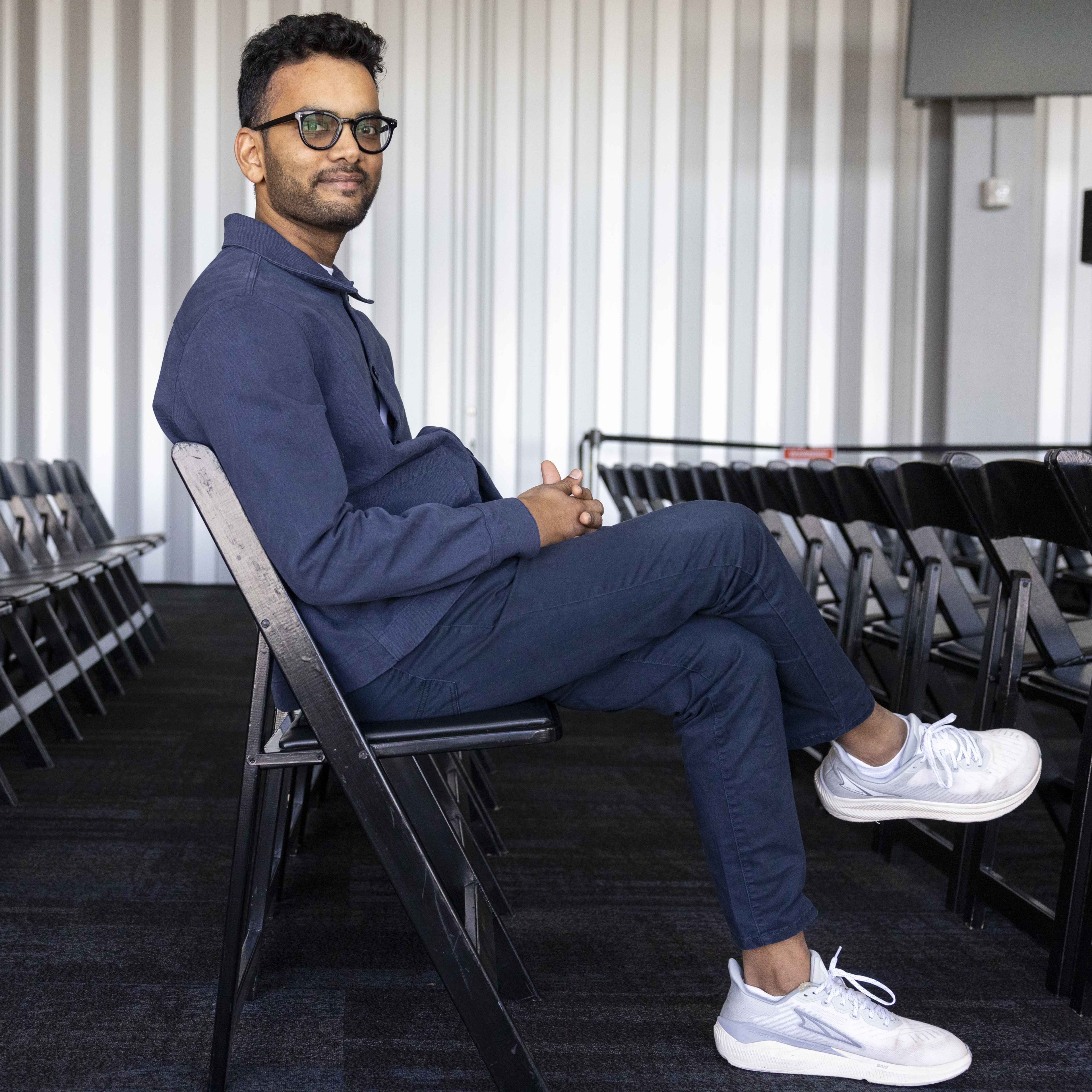 A man in glasses and a blue outfit sits cross-legged on a black chair, relaxed in a room with rows of empty chairs and a corrugated wall background.