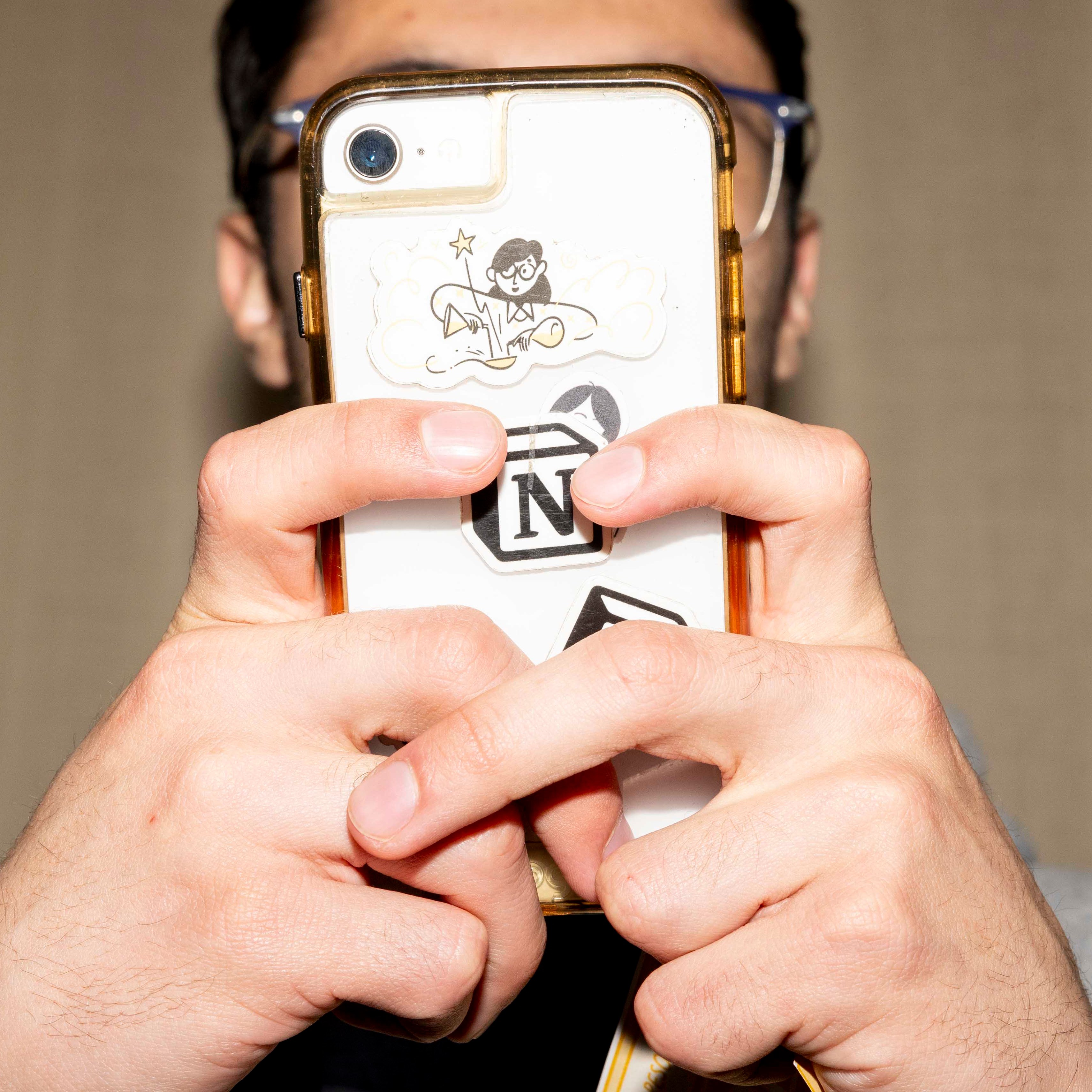 A person holds a phone with both hands, covering most of their face. The phone has a clear case displaying various playful stickers.