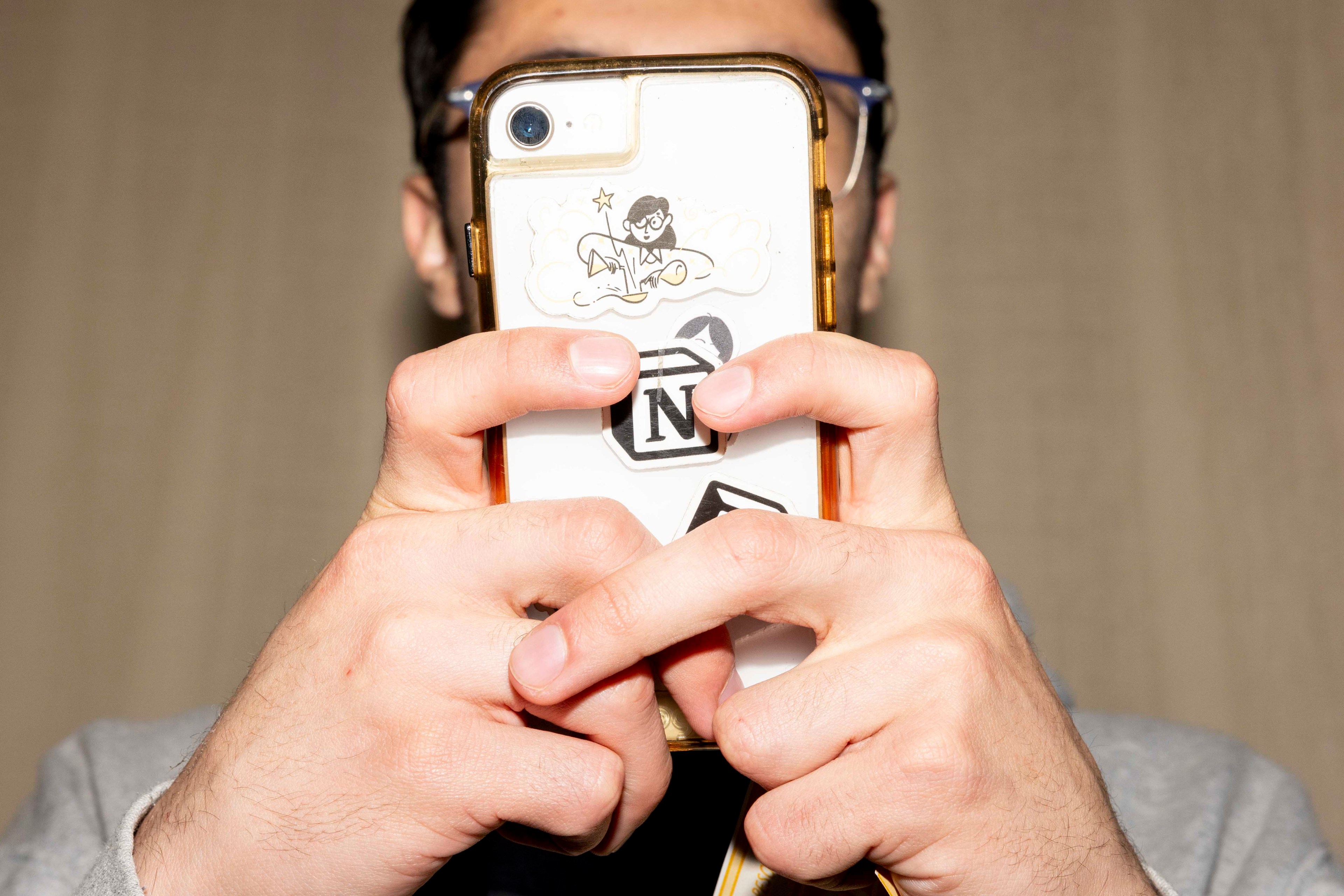 A person holds a phone with both hands, covering most of their face. The phone has a clear case displaying various playful stickers.