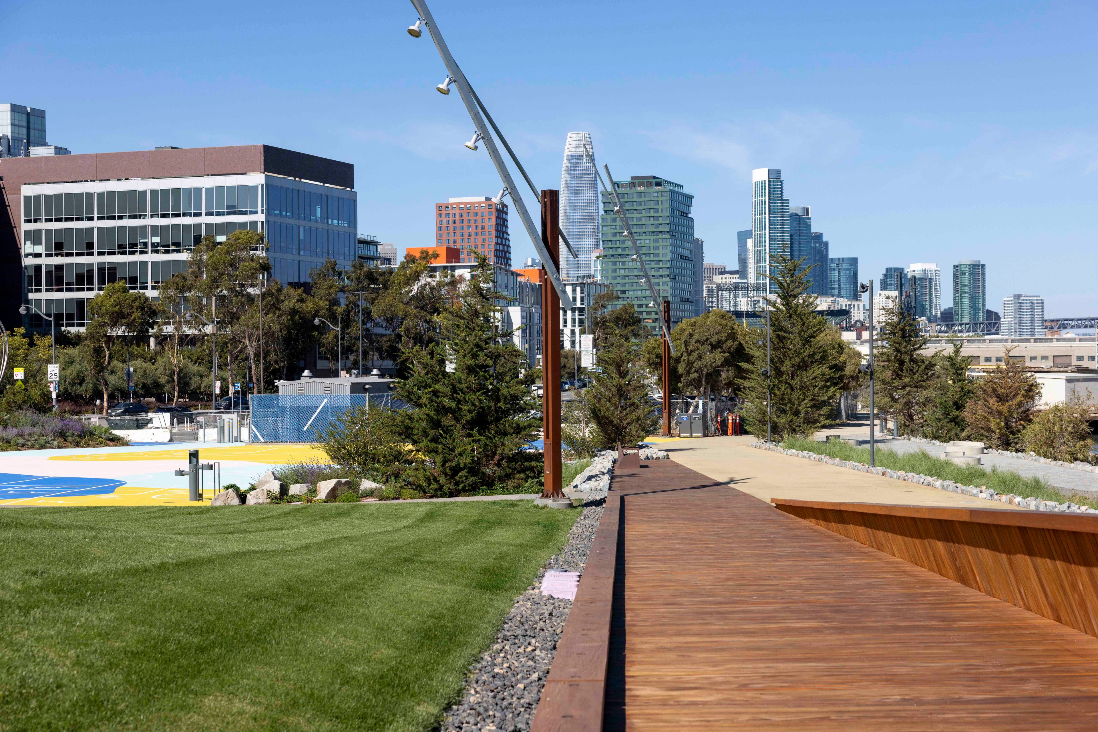 A paved walkway and a grassy area are lined with trees and modern buildings, set against a skyline of tall skyscrapers on a clear day.