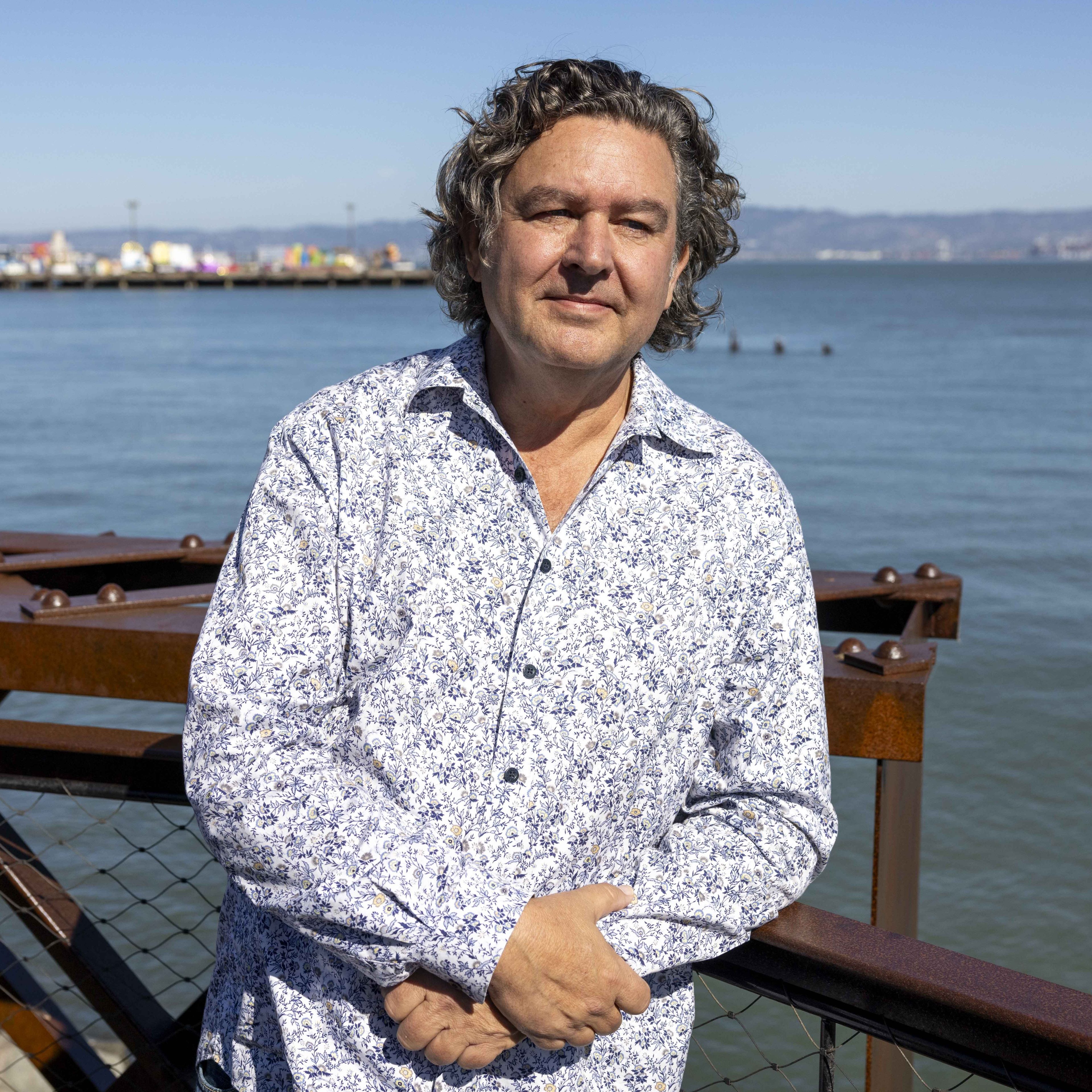 A person stands by a railing near a calm body of water. They are wearing a white and blue patterned shirt, and the background features a distant shoreline.