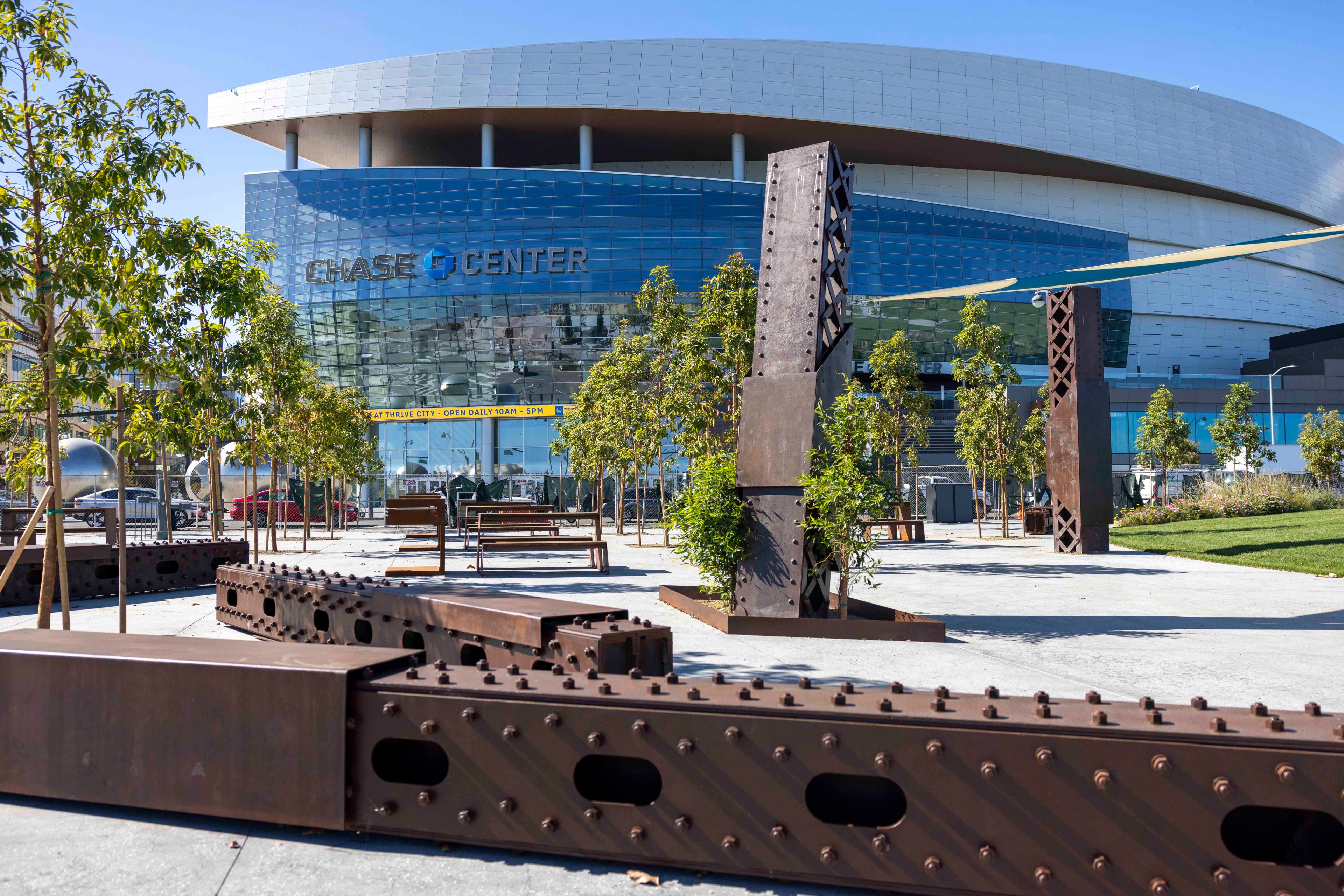 The image shows the Chase Center, a large, modern building with a curved, metallic facade. In the foreground are trees and abstract metal sculptures.