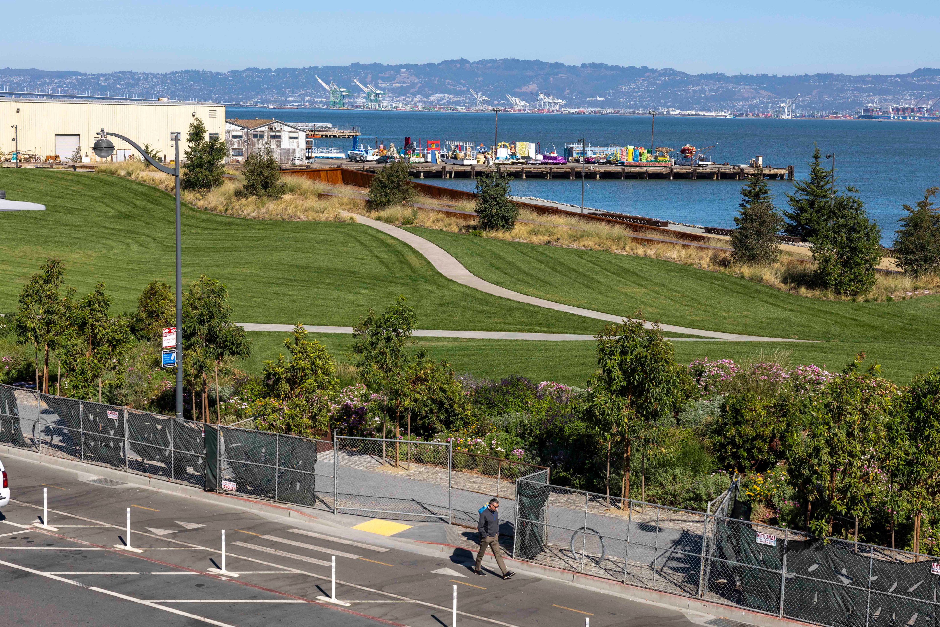 A green park by the waterfront features a winding path. Near the shore are industrial buildings and colorful containers. A person walks on the pavement.