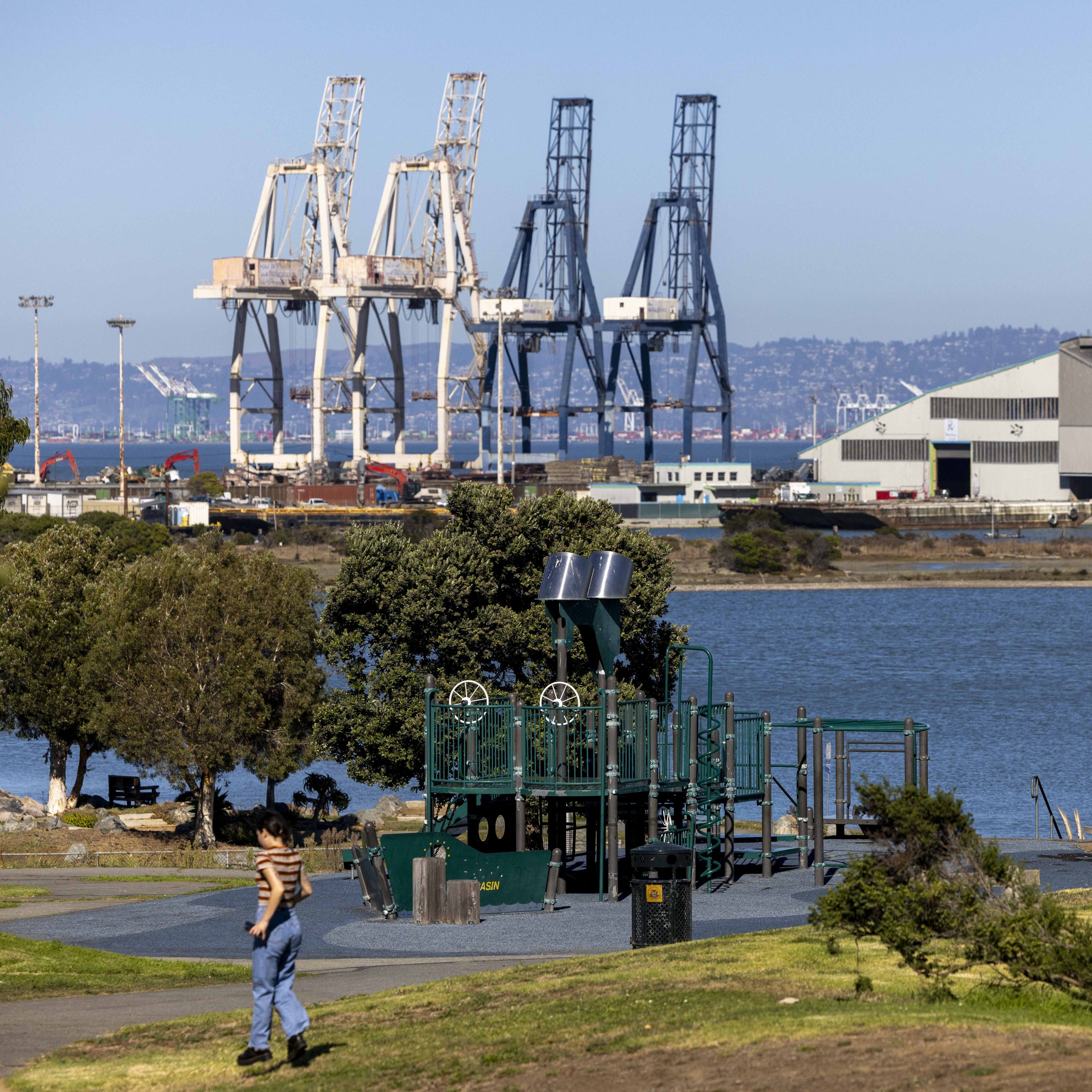 A park with a playground and a person walking in the foreground is set against a backdrop of cranes and industrial buildings by the water.