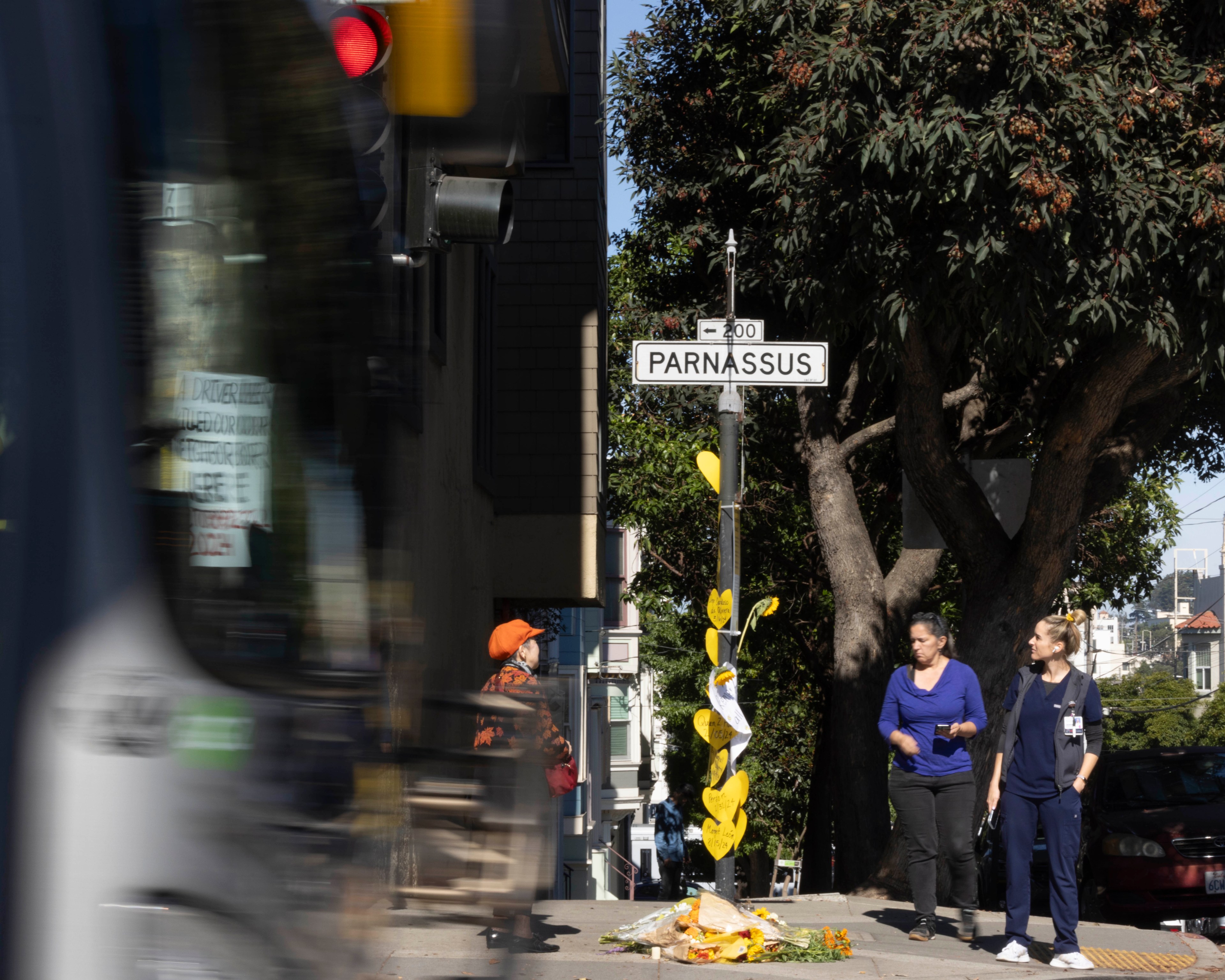 A street scene with a &quot;Parnassus&quot; sign, two people walking, and a tree. A blurred bus passes by, and there are yellow heart decorations and flowers on the post.