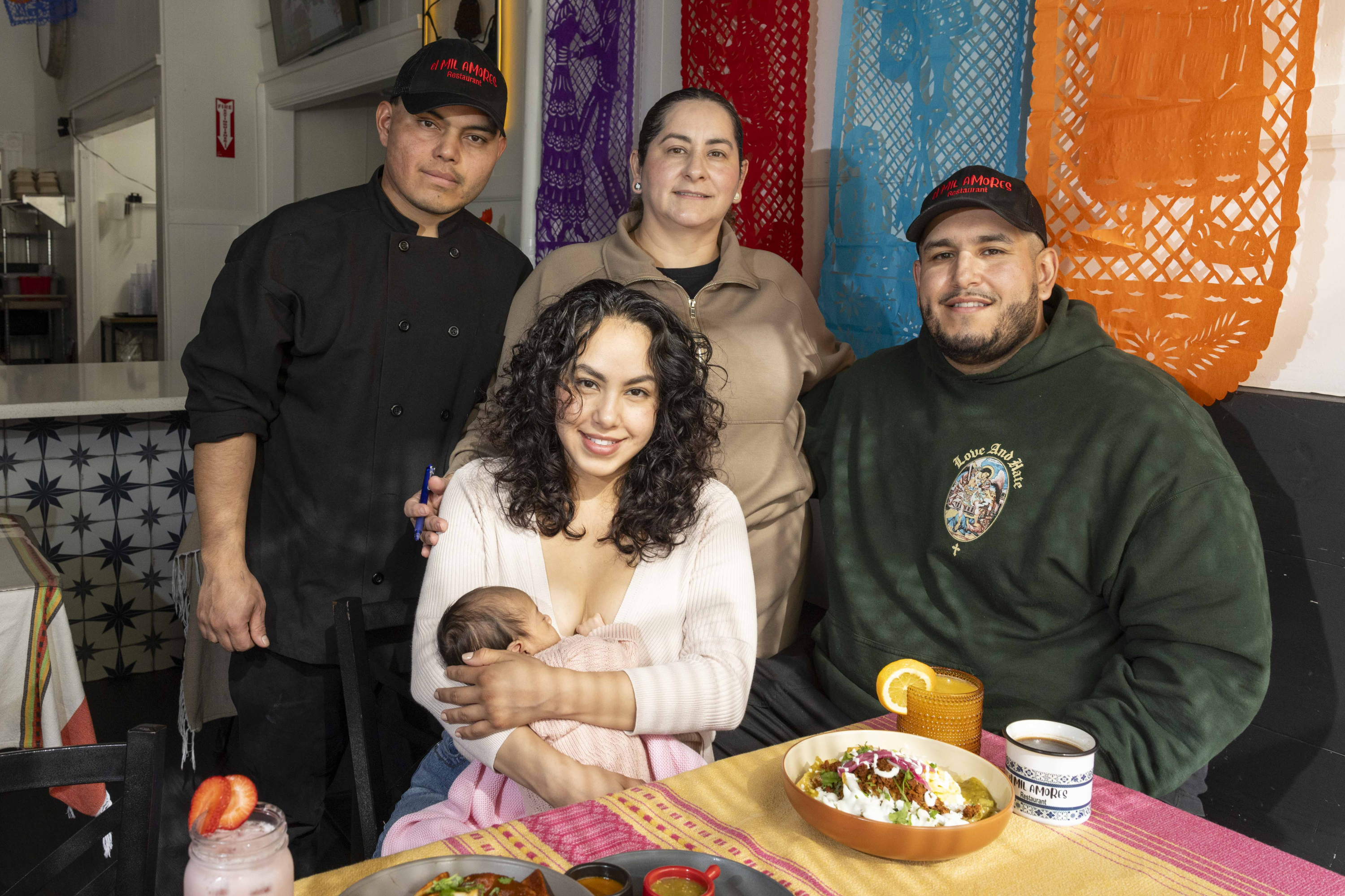 A group of four adults and a baby are in a colorful room with festive papel picado decorations. They're gathered around a table with food and drinks.