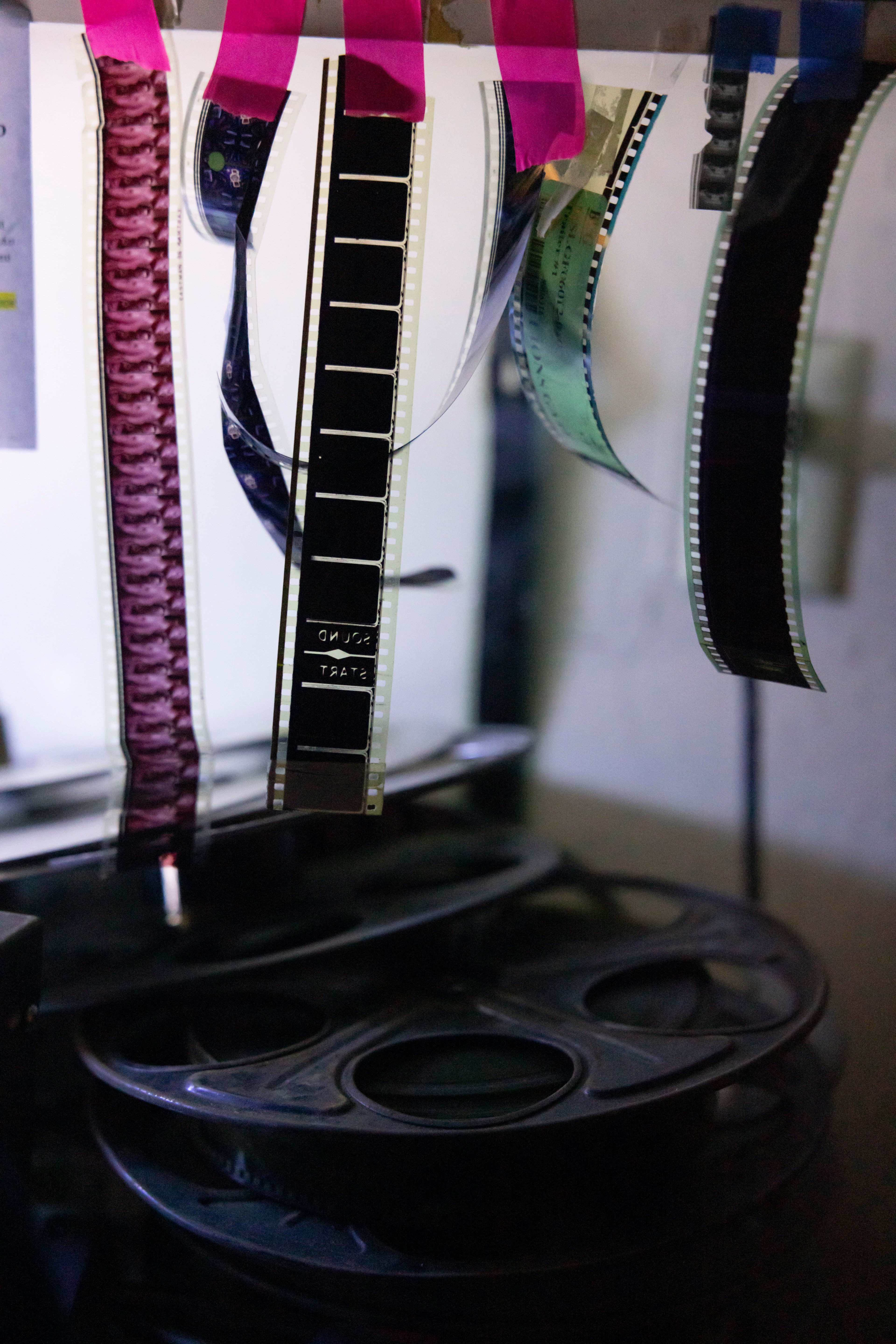 Strips of film hang from a shelf where they are attached with pink tape. Below them are horizontally placed reels of film.