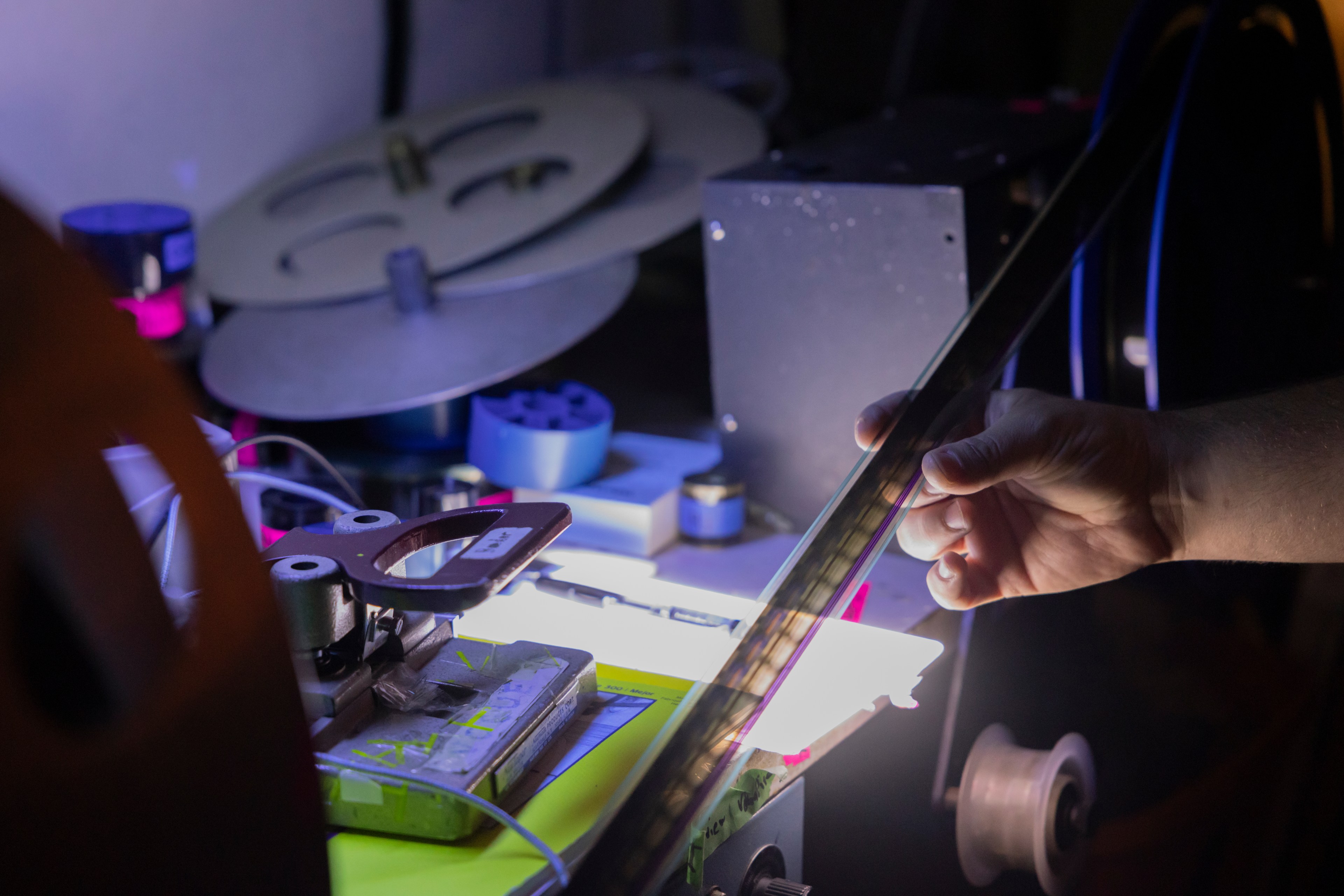 A person's hand holds a long strip of film steady as it is mechanically rolled back into a reel. Materials like parts of reels clutter the desk.