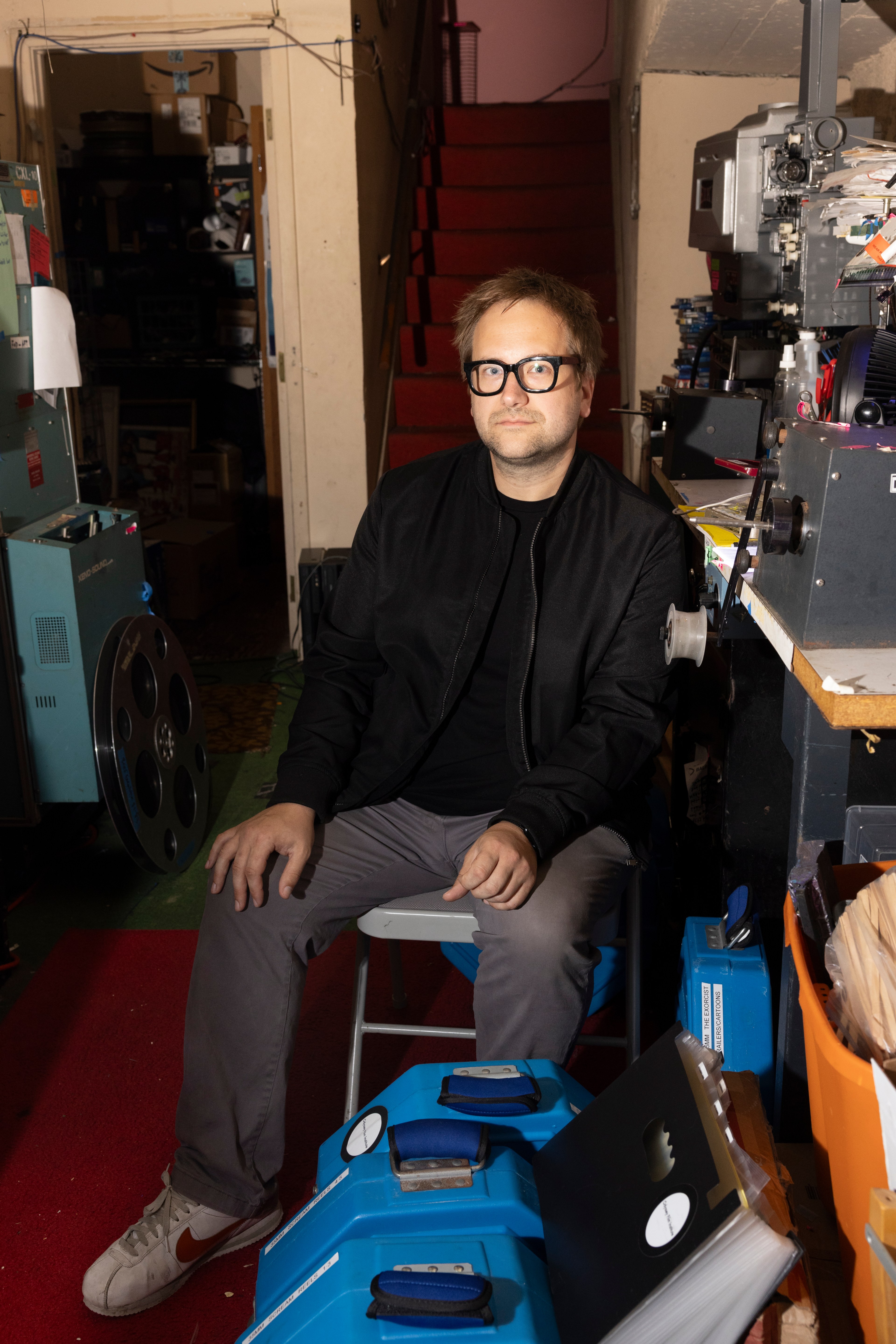 A man in a black shirt and jacket and gray pants sits facing the camera in a projection room surrounded by film reels and other materials.