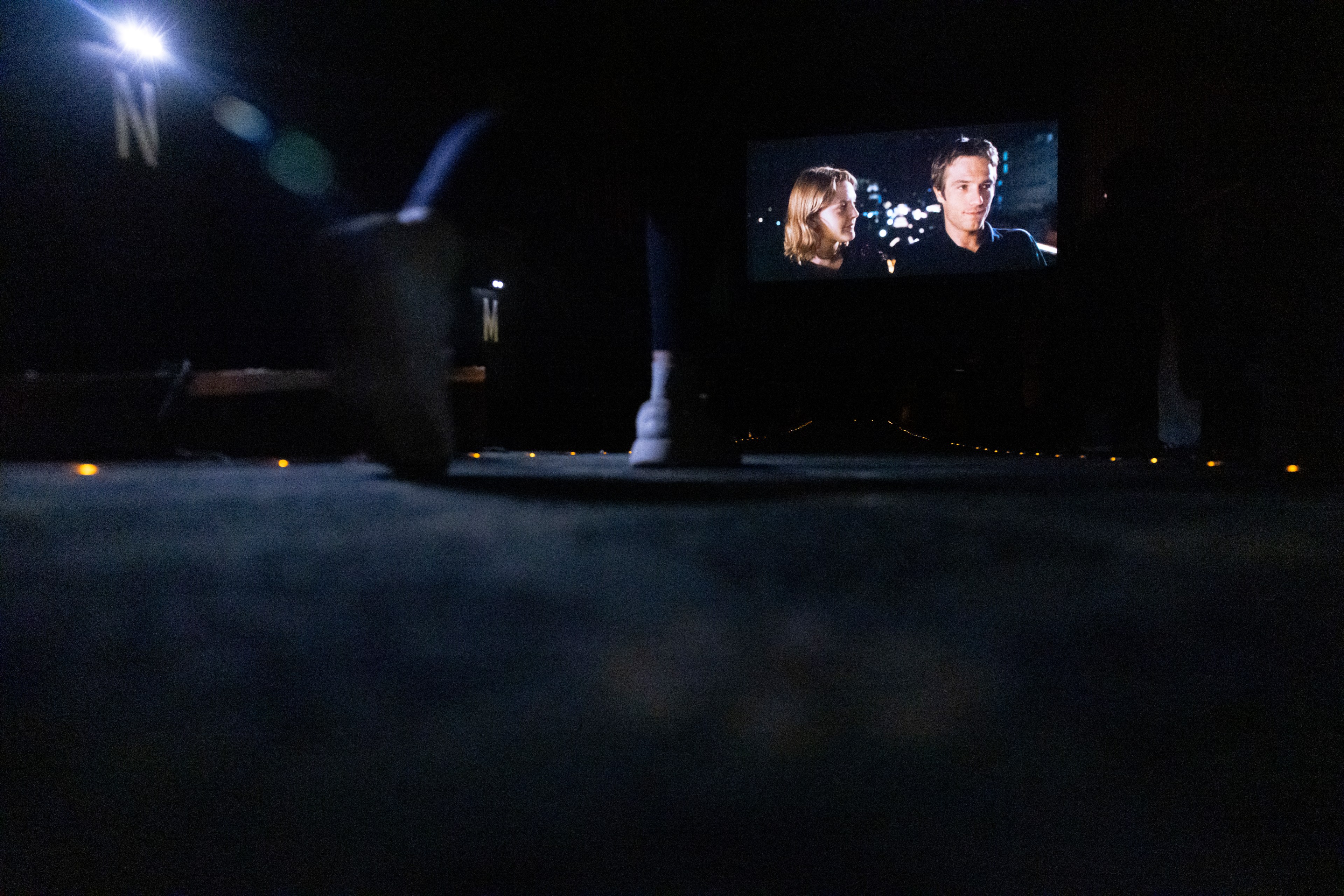 A person's feet are visible as they walk past rows of theater seats which have lights that illuminated the carpeted walkway. In the distance, a woman and a man are seen on a movie theater screen.