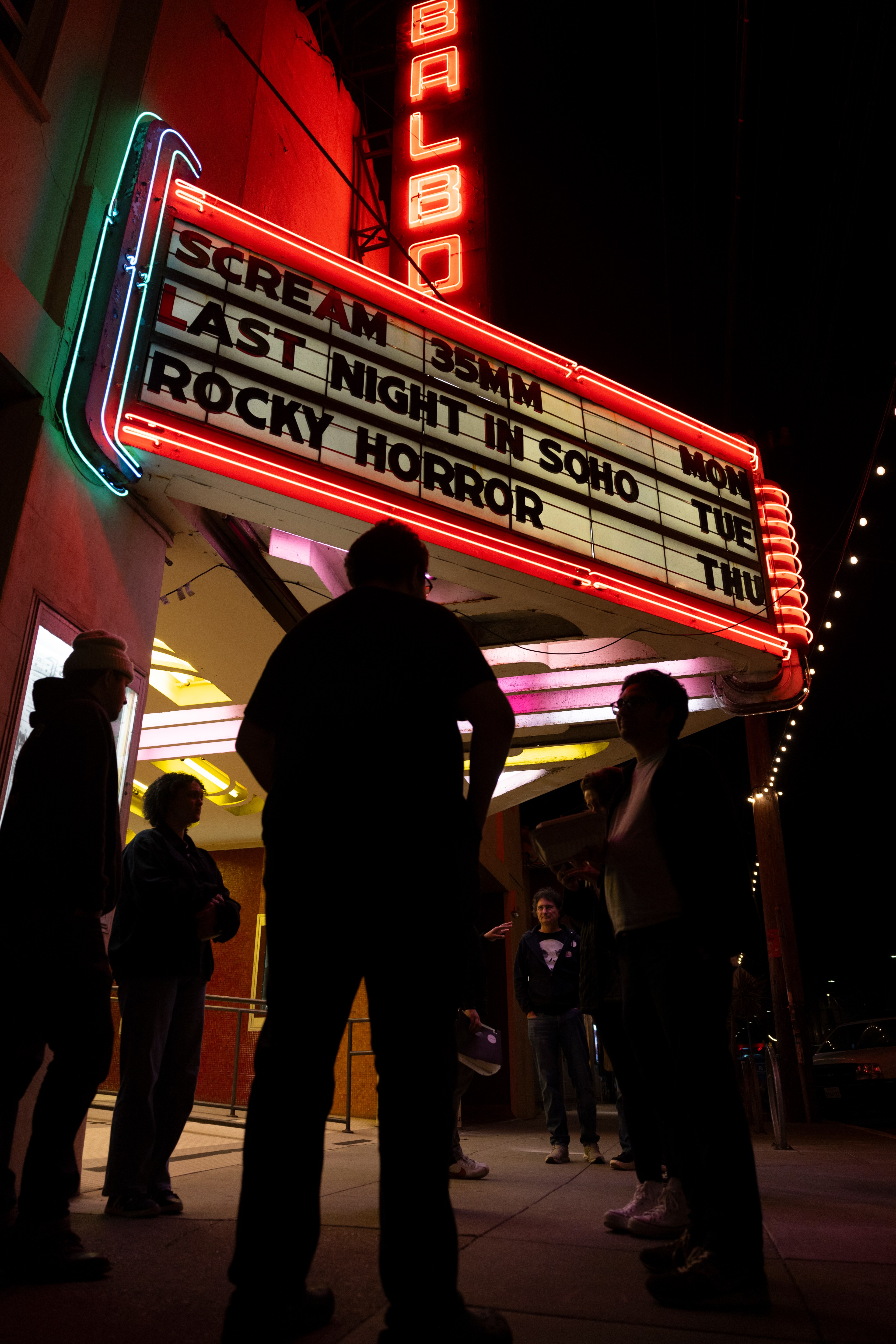 Silhouettes of people stand outside of a movie theater marquee with the name &quot;Balboa&quot; written vertically in lights above.