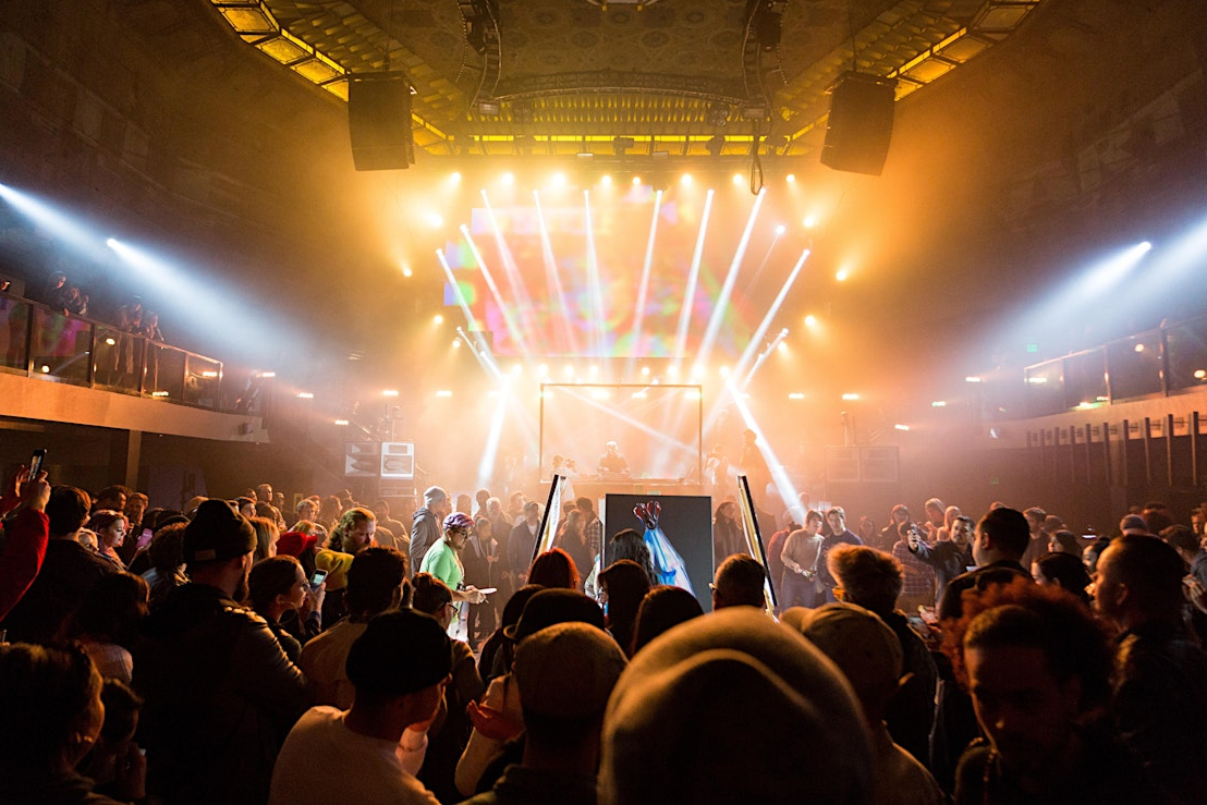 A vibrant concert scene shows a crowd gathered around a stage with dynamic lighting and colorful visuals, creating an energized atmosphere in a packed venue.