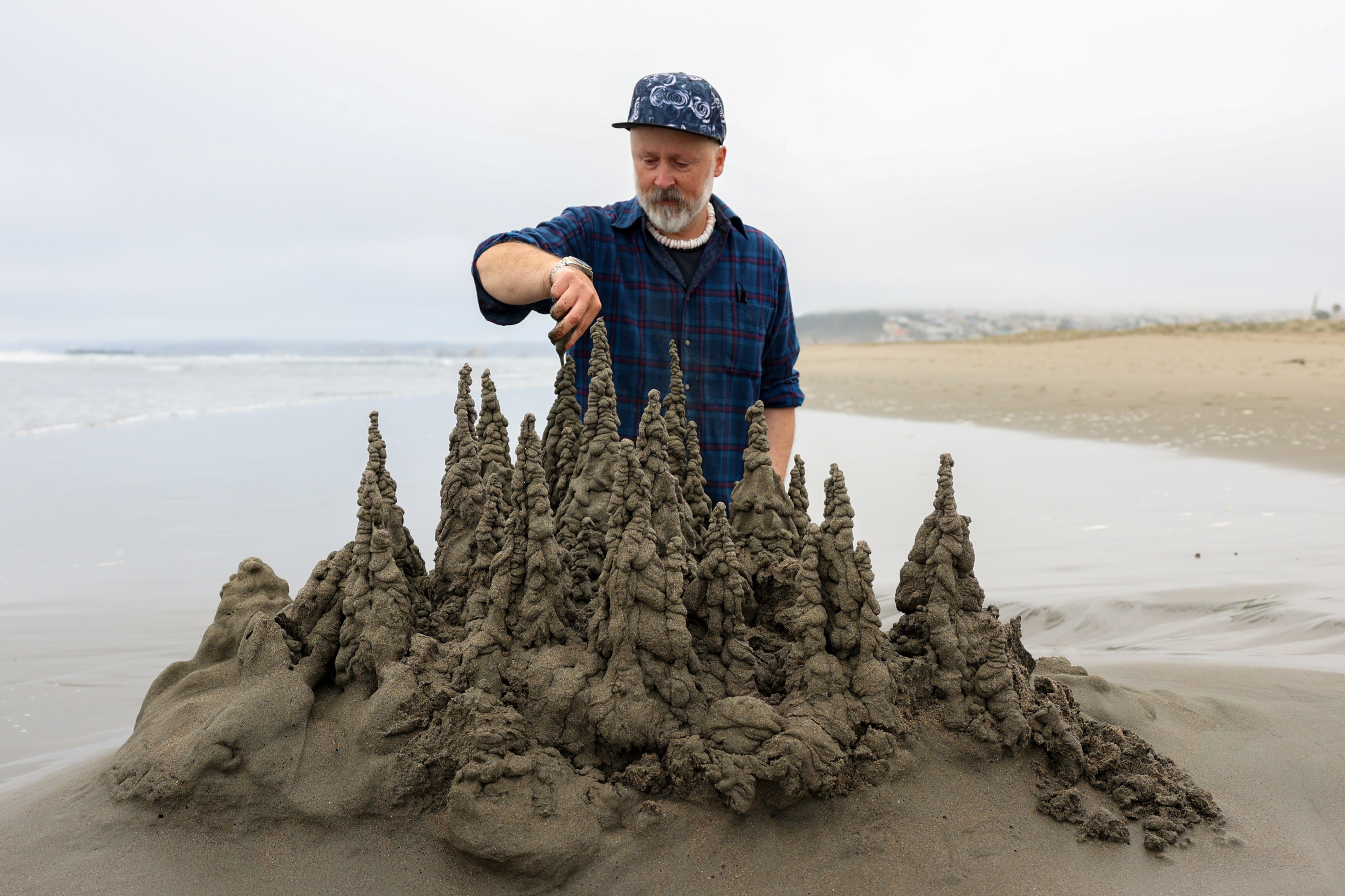 A person in a blue checkered shirt and a cap creates a intricate sandcastle with tall, spiky towers on a flat, overcast beach.