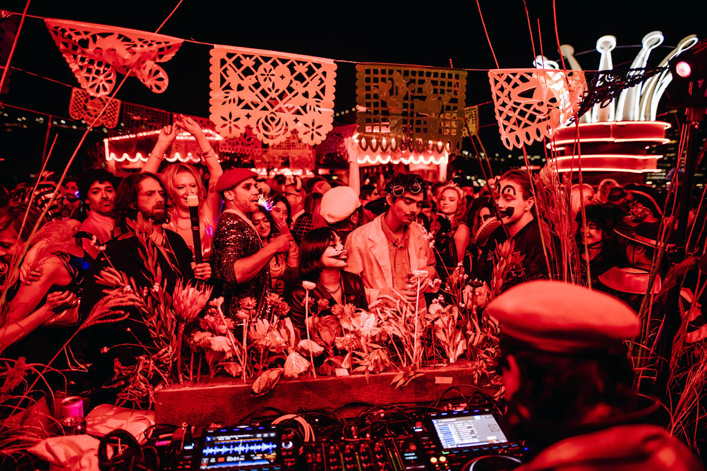 A lively party scene with people in costumes surrounded by vibrant red lighting. Decorative papers hang above, and a DJ setup features prominently in the foreground.