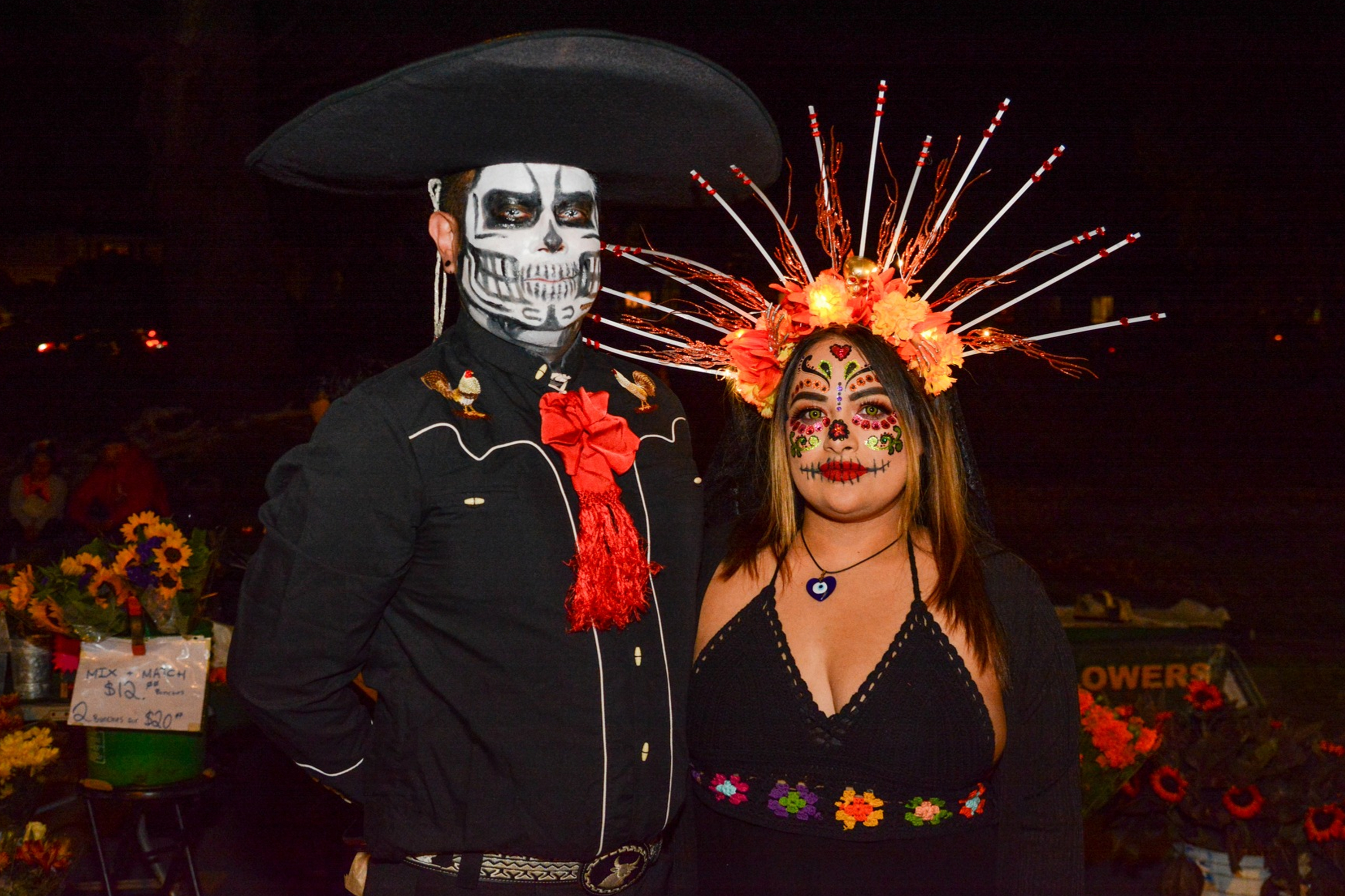 A couple in elaborate Day of the Dead costumes, with skeleton face paint and traditional attire. The woman wears a floral headdress, and the man has a decorative hat.