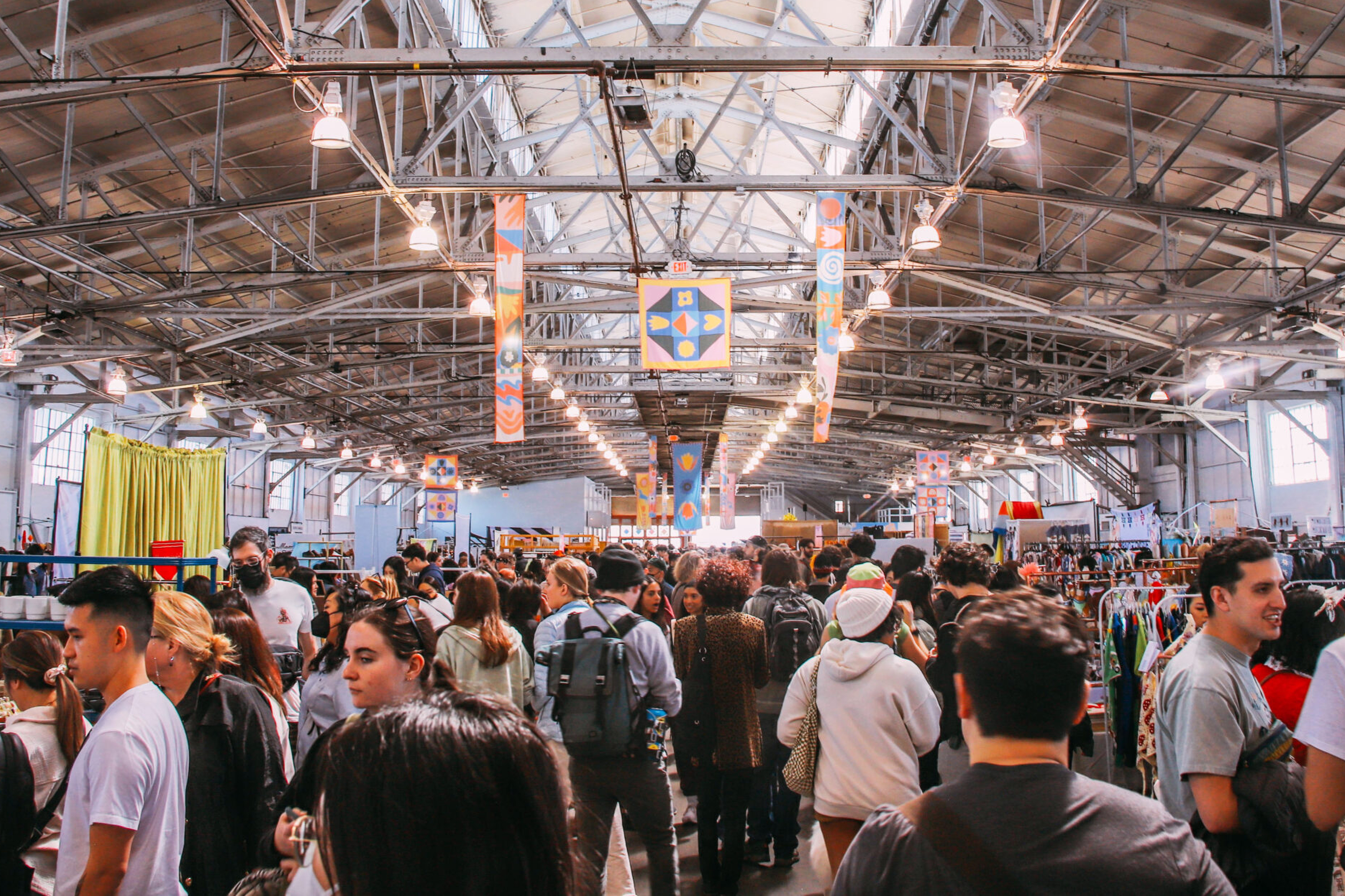 A bustling indoor market with many people mingling under a high, industrial-style ceiling adorned with colorful banners, surrounded by various stalls.