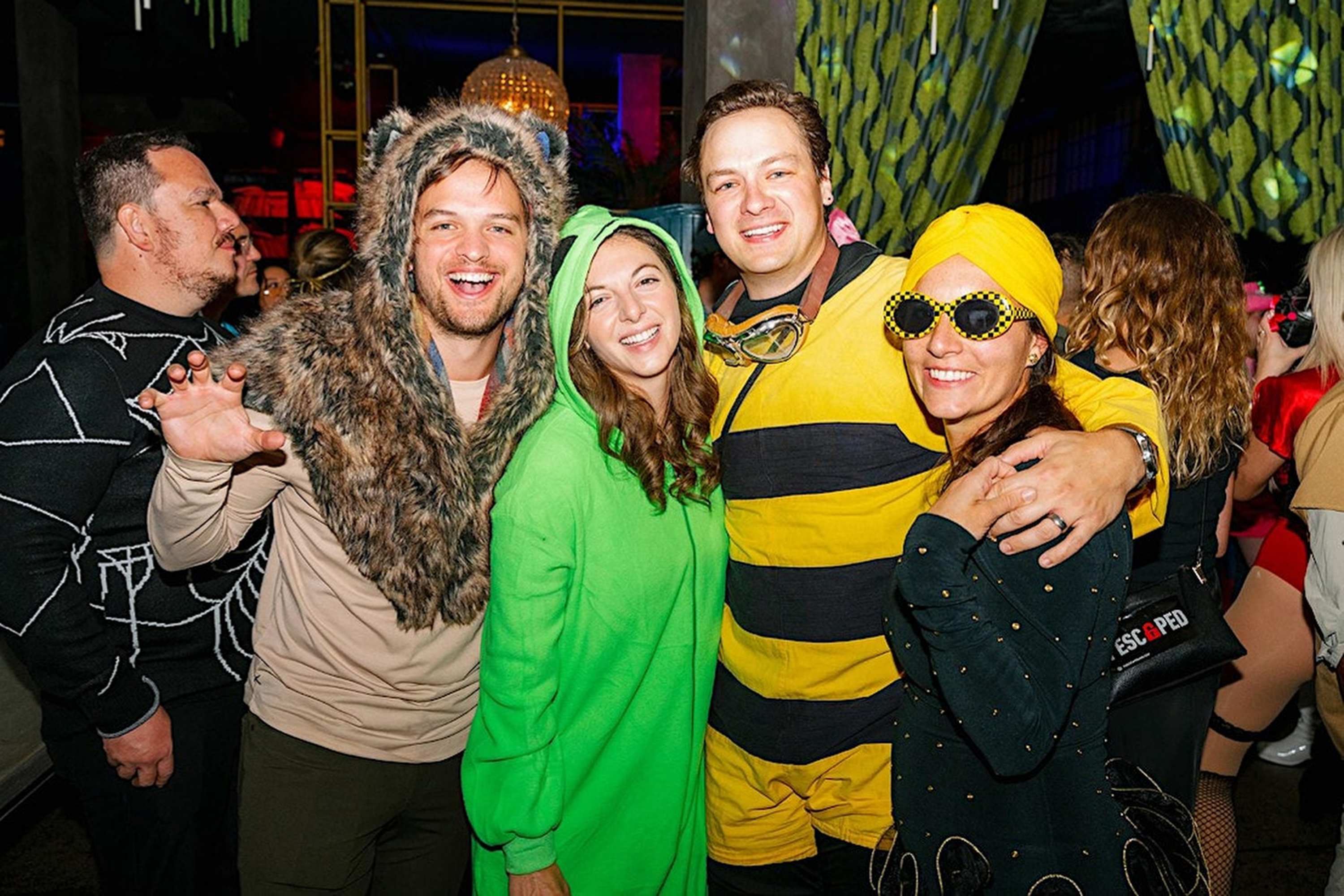 Four people in Halloween costumes pose for a photo. One is in a wolf costume, another in green, one as a bee, and the last with yellow accessories and polka dots.