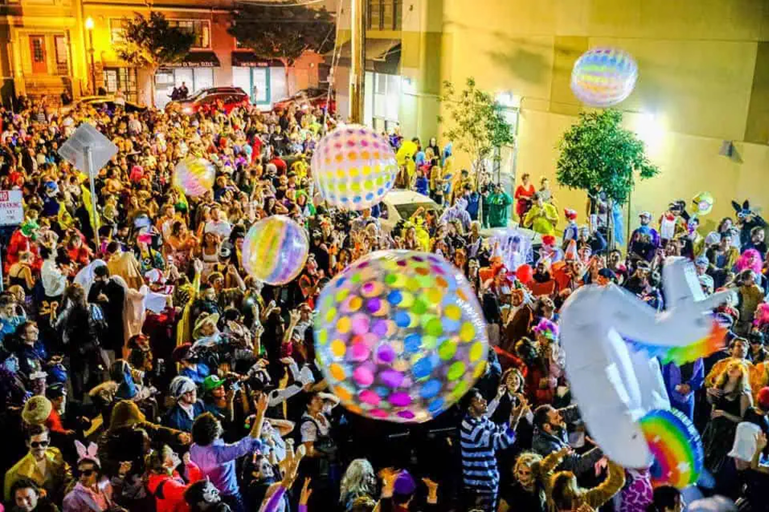 A lively street festival features a large crowd in colorful costumes and hats, surrounded by several large, multicolored inflatable balls being tossed around.