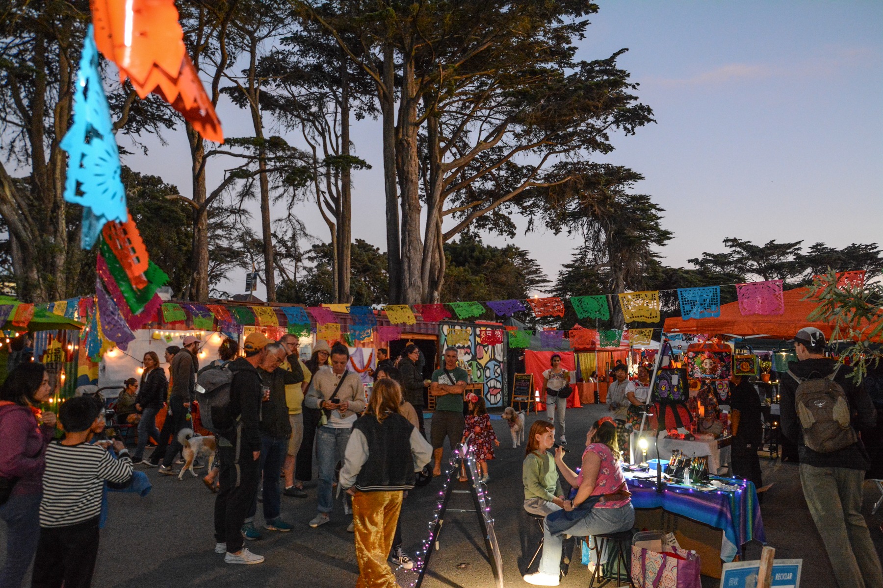 A lively outdoor festival with colorful flags hanging above. People gather around various stalls, and one person is getting their face painted. Tall trees surround the scene.