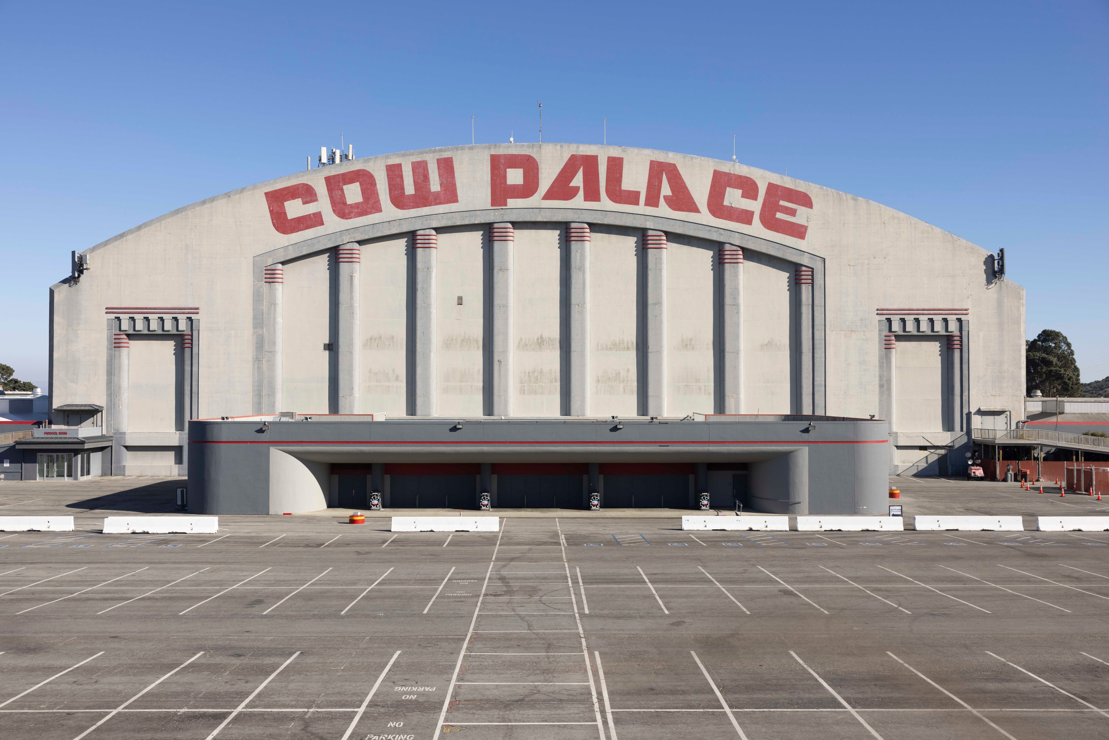 A large, grey building with &quot;COW PALACE&quot; in red letters, featuring an empty parking lot and a clear, blue sky above.