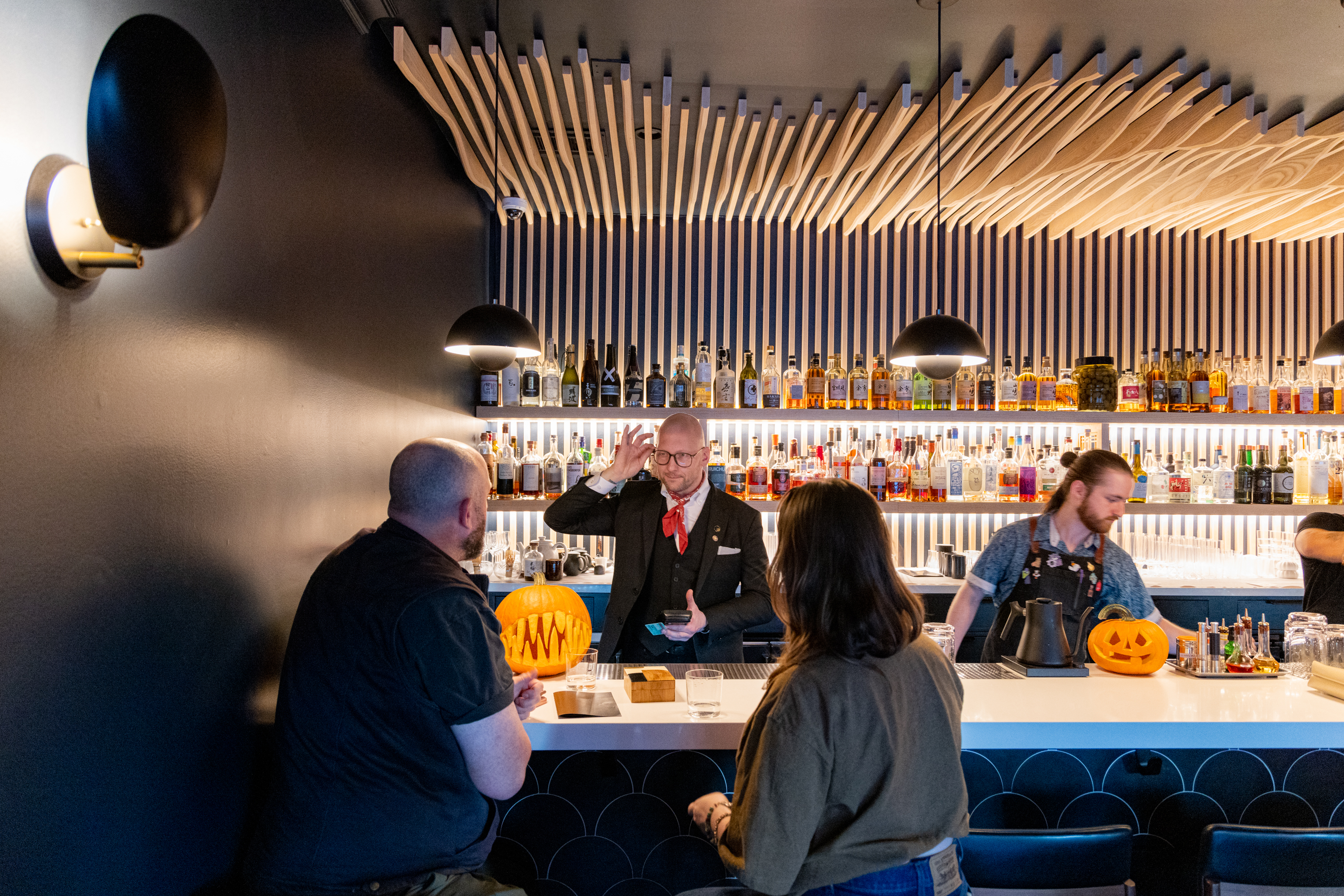 A bartender gestures while talking to three customers at a bar with a modern design. Shelves of liquor bottles and a carved pumpkin are visible behind them.