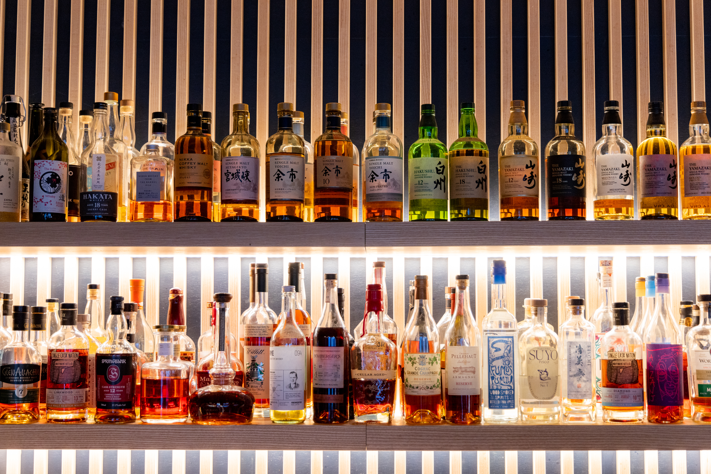 The image shows two shelves filled with various liquor bottles, mainly whiskies, with diverse labels and colors, set against a wooden slatted backdrop.