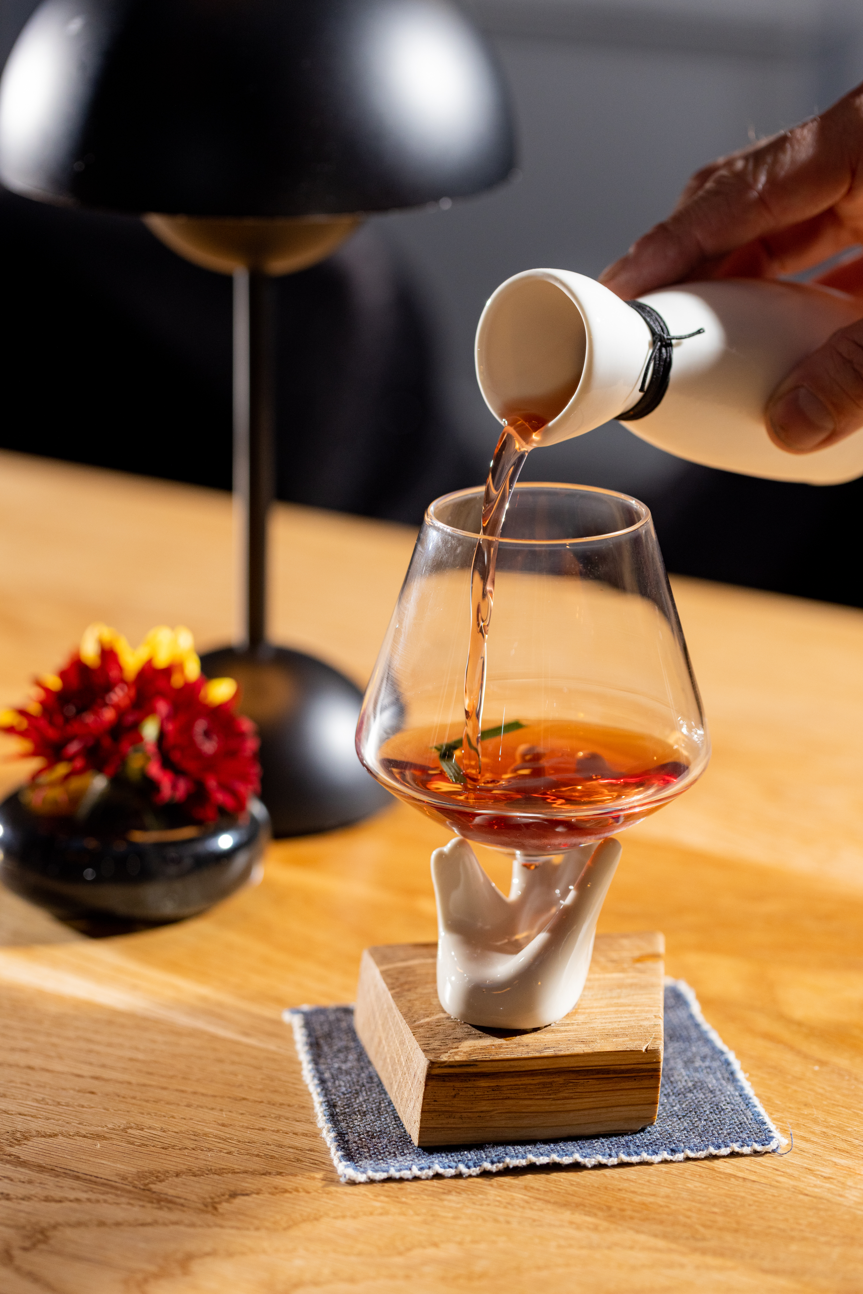 A hand-shaped holder supports a glass being filled with liquid from a small white pitcher, set on a wooden base. A black lamp and red flowers are in the background.