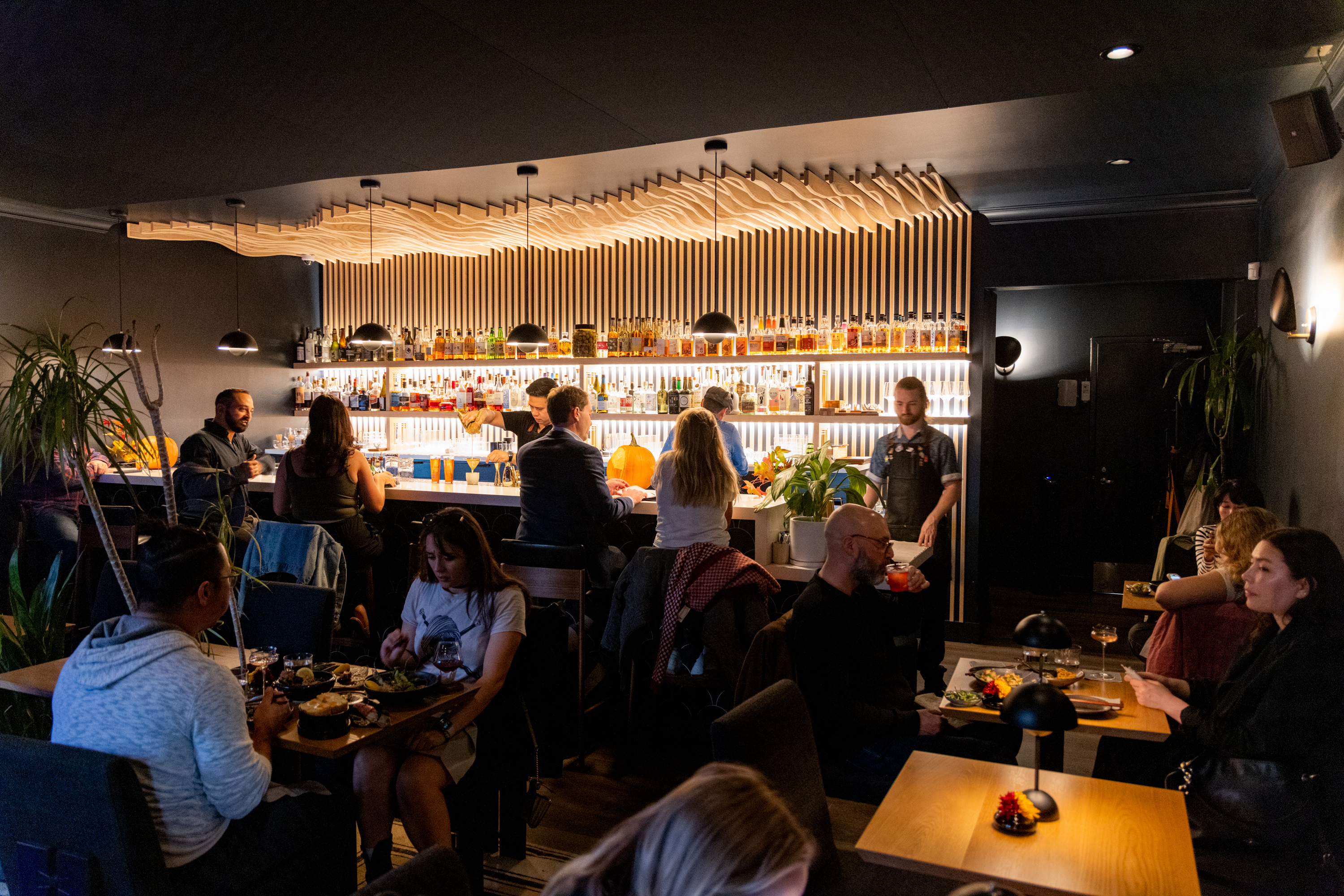 A cozy bar scene with people dining and chatting. A well-lit bar area serves drinks, with bottles neatly displayed on shelves. The atmosphere is lively and inviting.