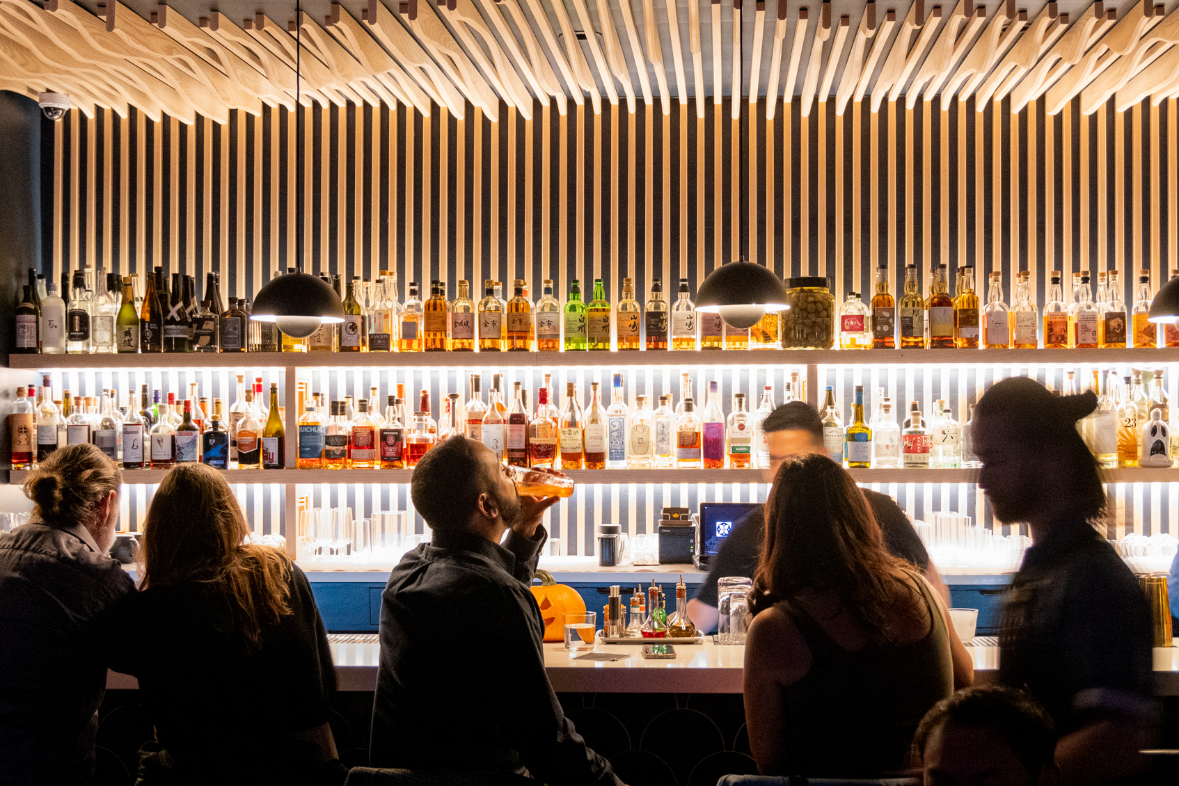 A bar with backlit shelves filled with bottles, featuring several people sitting and standing, and a bartender preparing drinks under modern pendant lights.