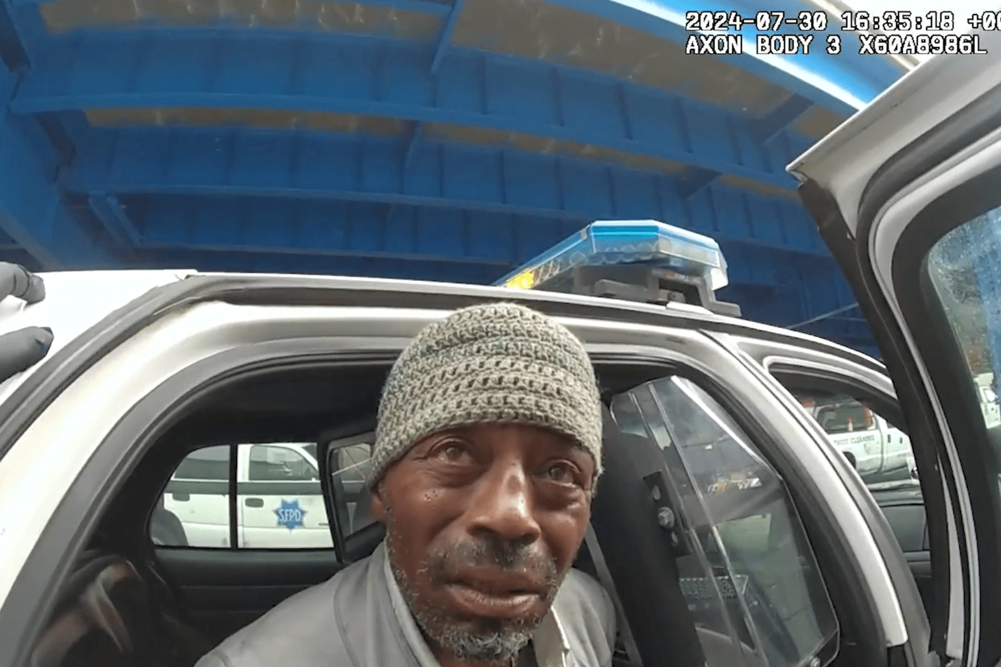 A man wearing a knit cap sits in the back of a police car with the door open. A gloved hand appears on the door frame, and a blue structure is visible above.