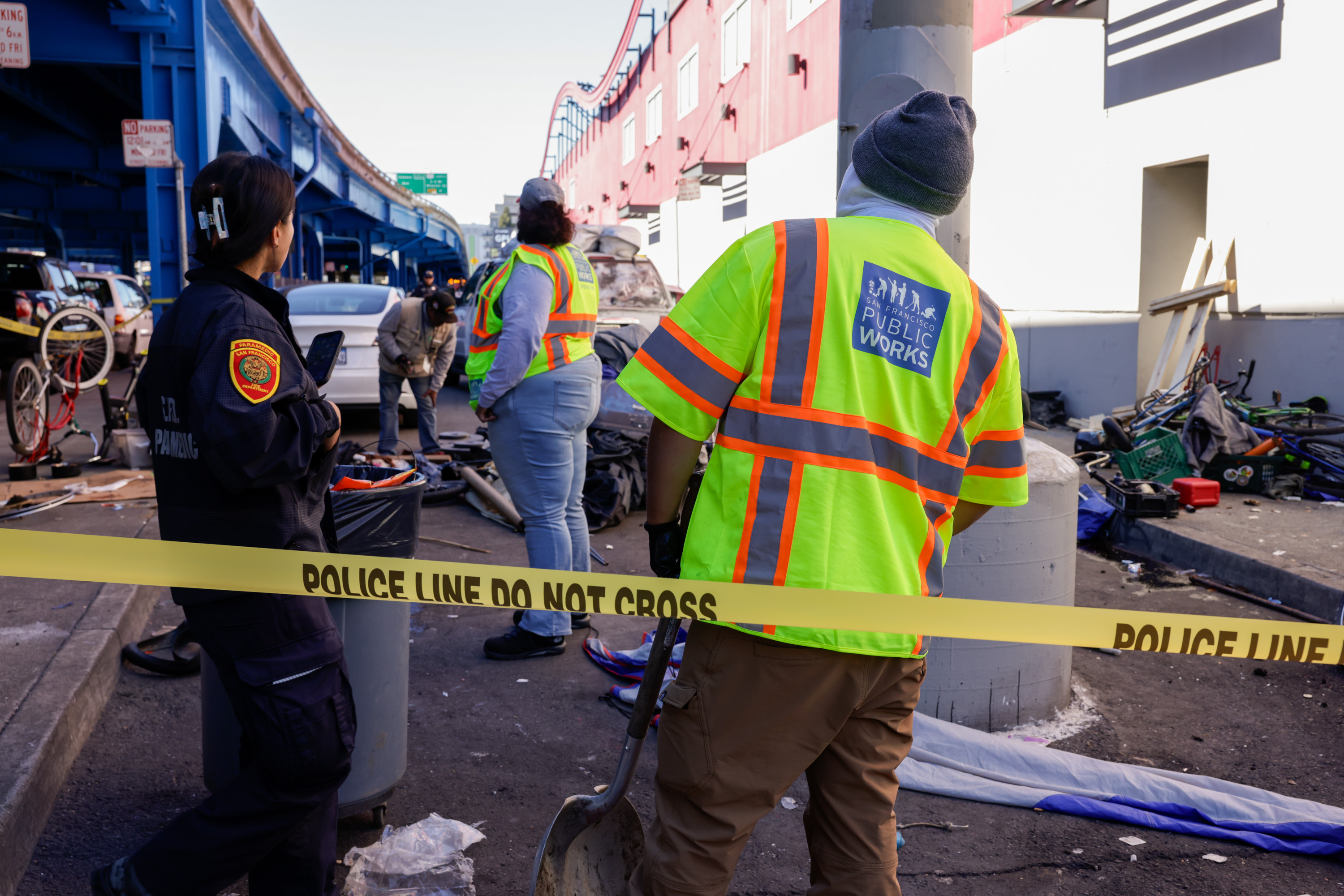 Several people are working near a busy street, with one in a public works vest. A police line is set up, and there's debris and bicycles around.